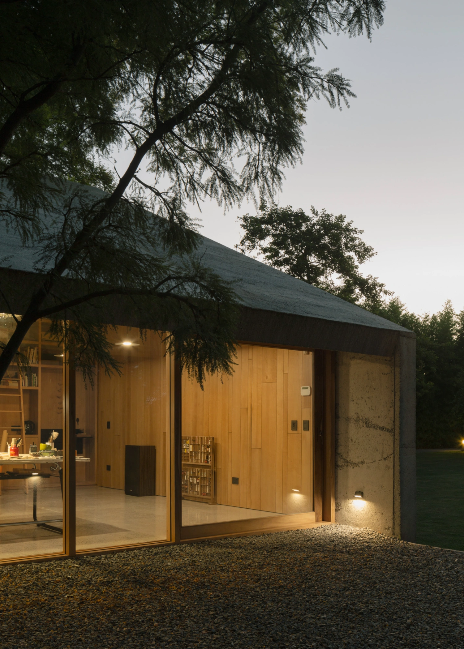 Atelier Workshop by Agustín Berzero — Evening photograph captures the workshop's southern glazed elevation as the primary focal point, its timber-framed glass strip glowing warm against the...