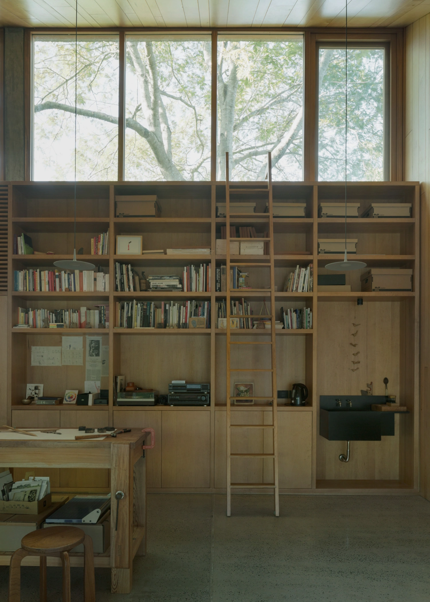 Atelier Workshop by Agustín Berzero — Interior view of the workshop revealing a wall-length timber shelving system with glass doors, organized across multiple bays by a central ladder.