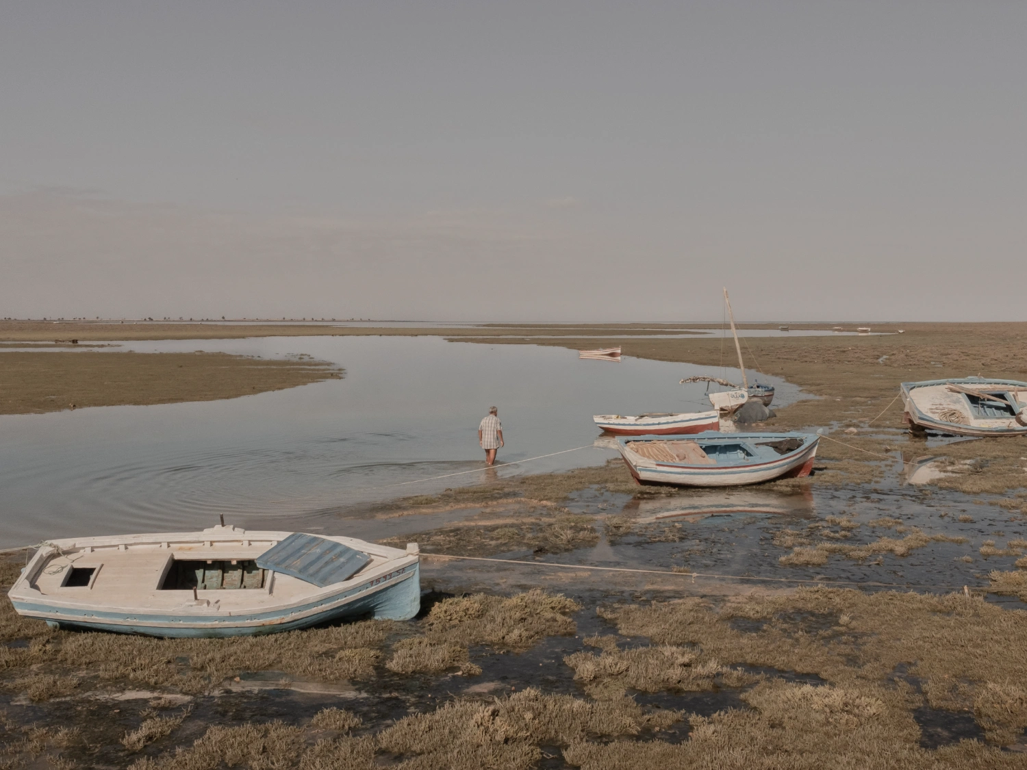 Where Dust and Water Dream Together by Skander Khlif — Boats rest on exposed mudflats in a tidal estuary, their hulls settled into dried earth and shallow water channels.