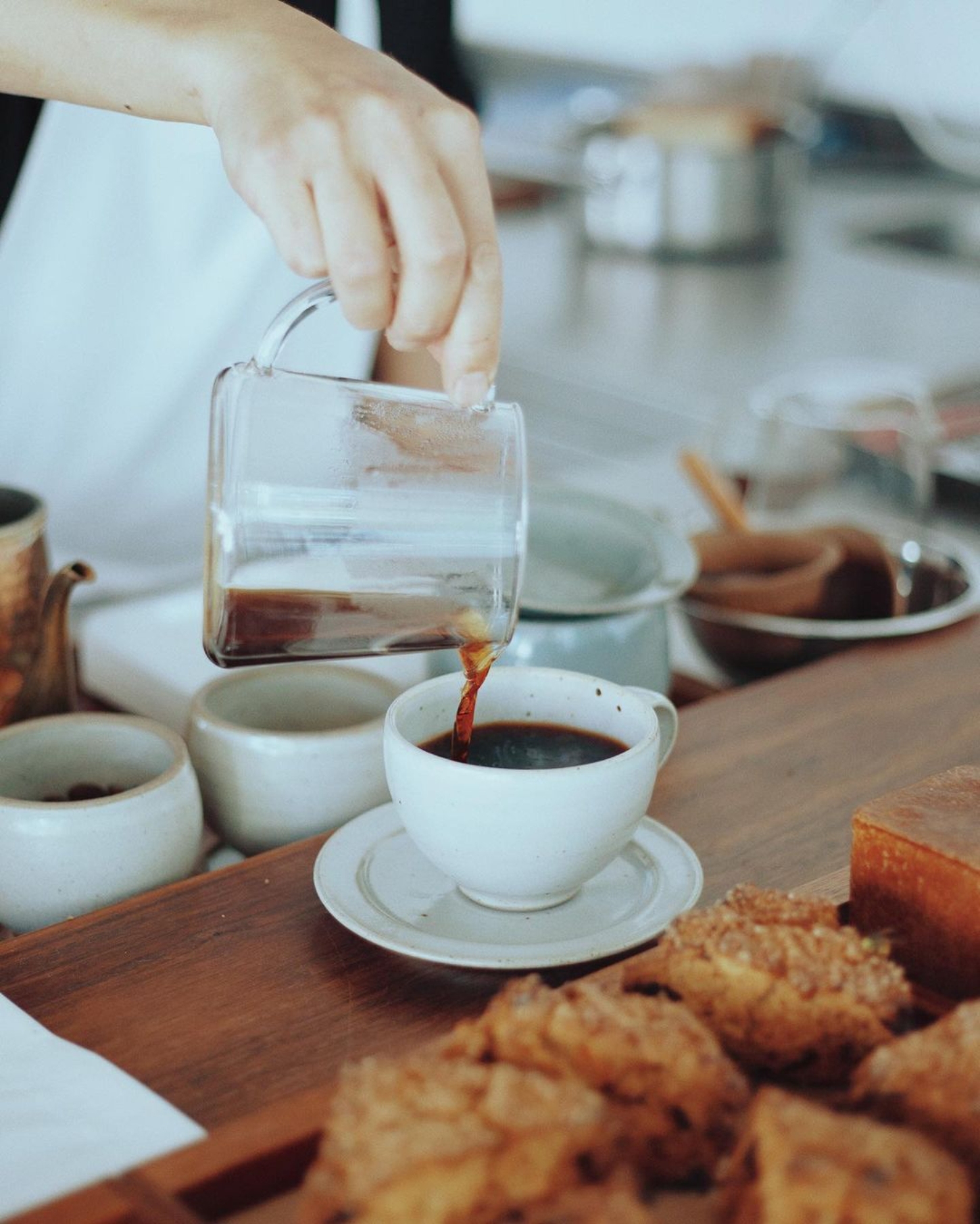 Julius in Berlin — A hand pours dark liquid from a glass vessel into a white ceramic cup on a saucer, captured mid-pour against a wooden table surface.
