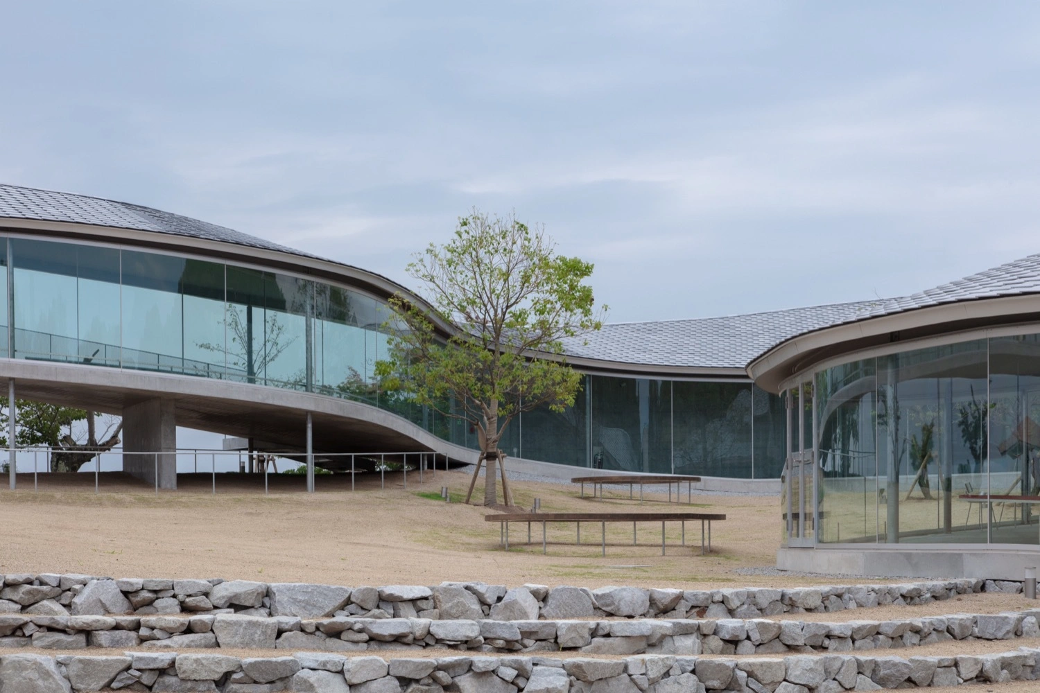 Yashima Mountaintop Park by SUO + Style-A — The mountaintop pavilion reveals itself as a permeable concrete ribbon, its curved form wrapping around a sandy courtyard anchored by a single tree.