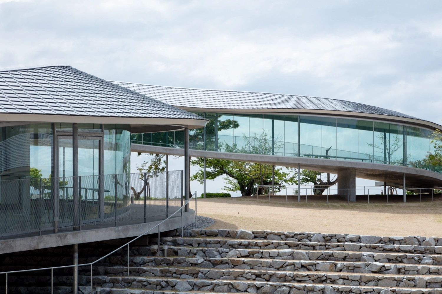 Yashima Mountaintop Park by SUO + Style-A — The mountaintop pavilion reveals its permeable threshold condition in this view, where a continuous white concrete ribbon curves around an expansive sandy...