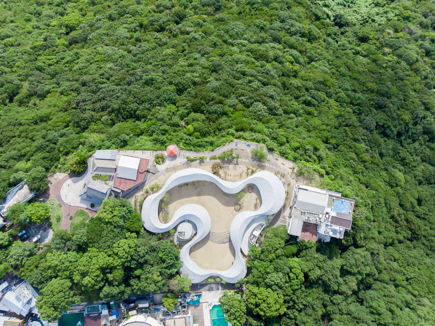 Yashima Mountaintop Park by SUO + Style-A — Aerial view of Yashima Mountaintop Park reveals a continuous white concrete ribbon structure wrapping around organic, undulating voids that expose sandy...