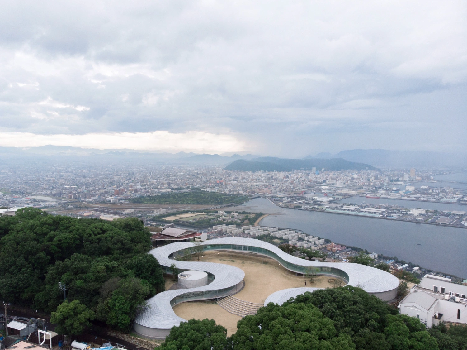 Yashima Mountaintop Park by SUO + Style-A — Aerial view of Yashima Mountaintop Park reveals the white concrete ribbon's full topographic sweep from above, its flowing geometry unspooling across the...