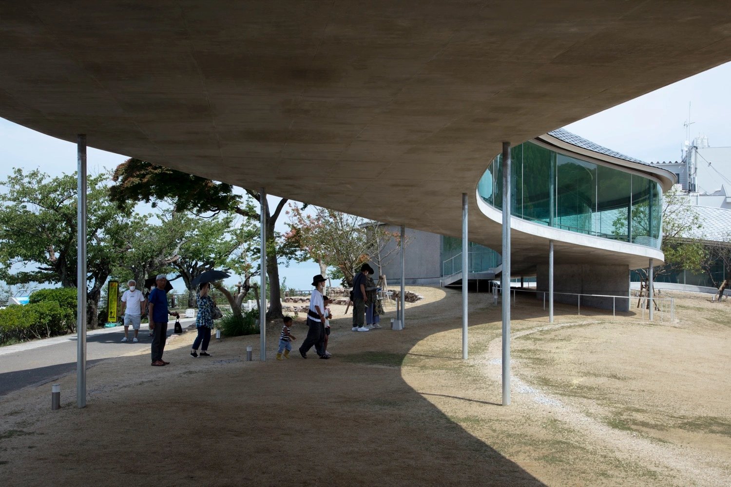 Yashima Mountaintop Park by SUO + Style-A — Visitors circulate beneath a continuous white concrete canopy that hovers above sandy ground, its underside a monolithic plane supported by slender steel...