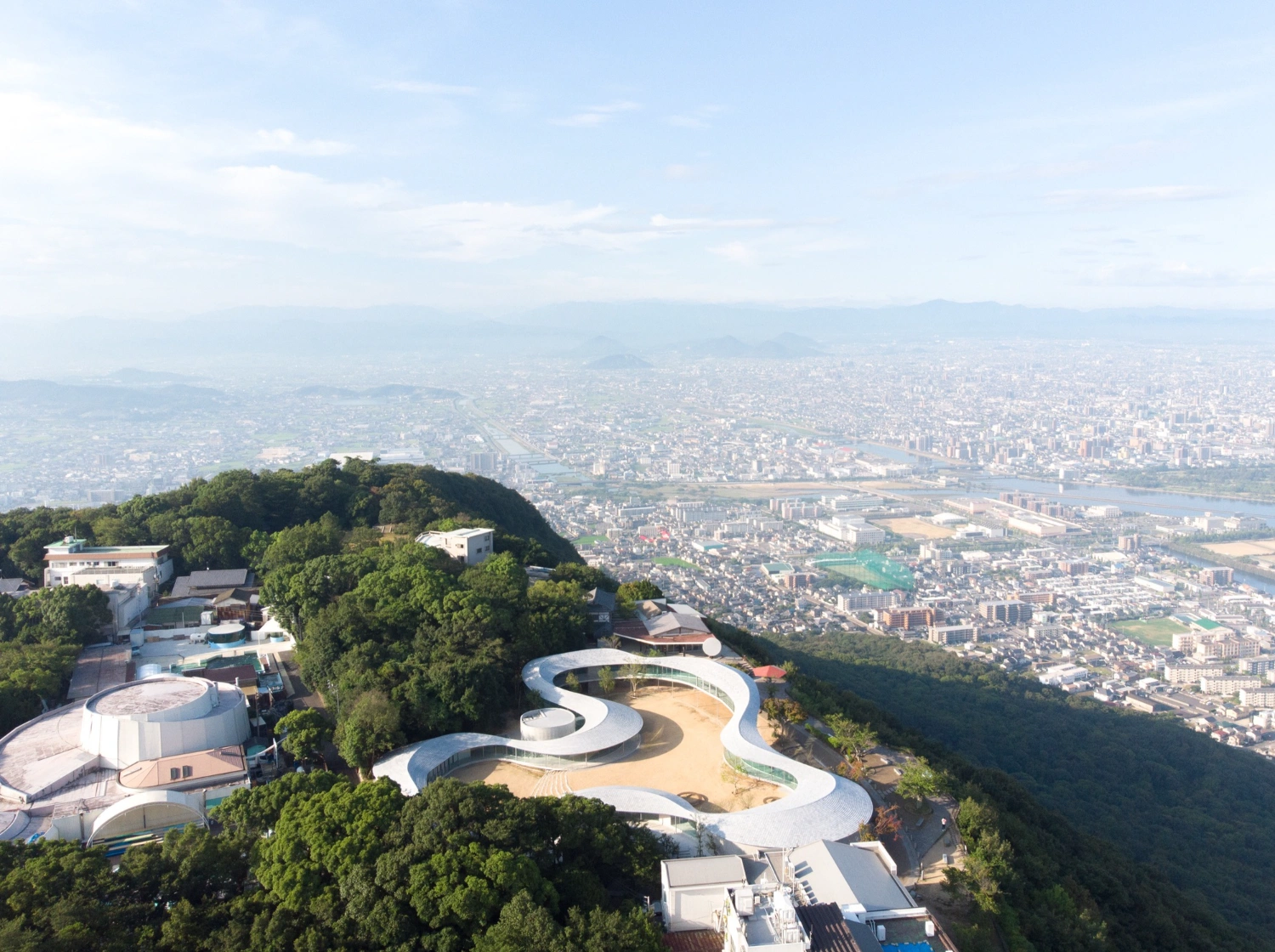 Yashima Mountaintop Park by SUO + Style-A — Aerial view of Yashima Mountaintop Park reveals a continuous white concrete ribbon structure wrapping around sandy interior courtyards, positioned on a...