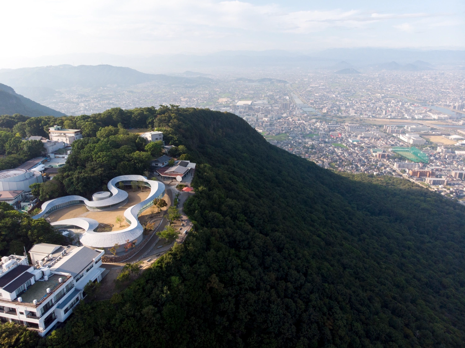 Yashima Mountaintop Park by SUO + Style-A — Aerial view of the Yashima Mountaintop Park revealing the white concrete ribbon structure winding across the forested slope, with a circular void element...