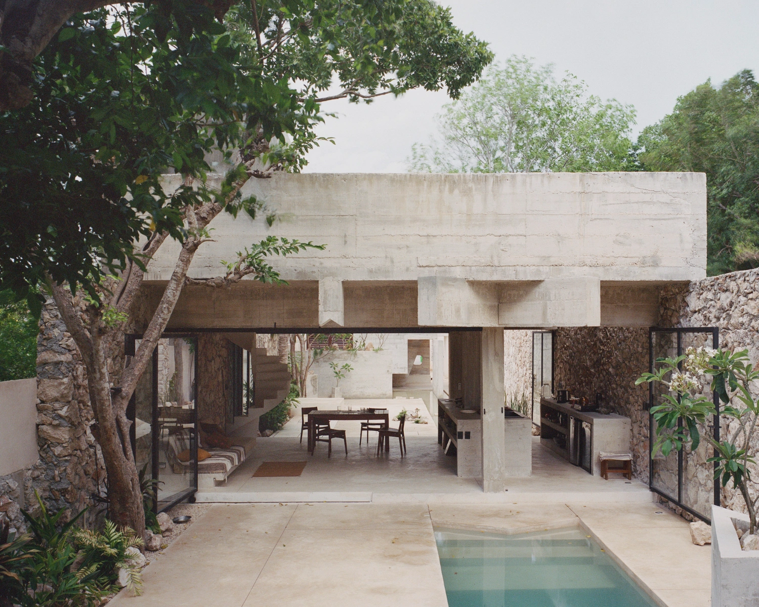 House in Los Cocos by Ludwig Godefroy Architecture — A view through the open-plan pavilion toward the courtyard, where a rectangular water basin with planted island anchors the composition.