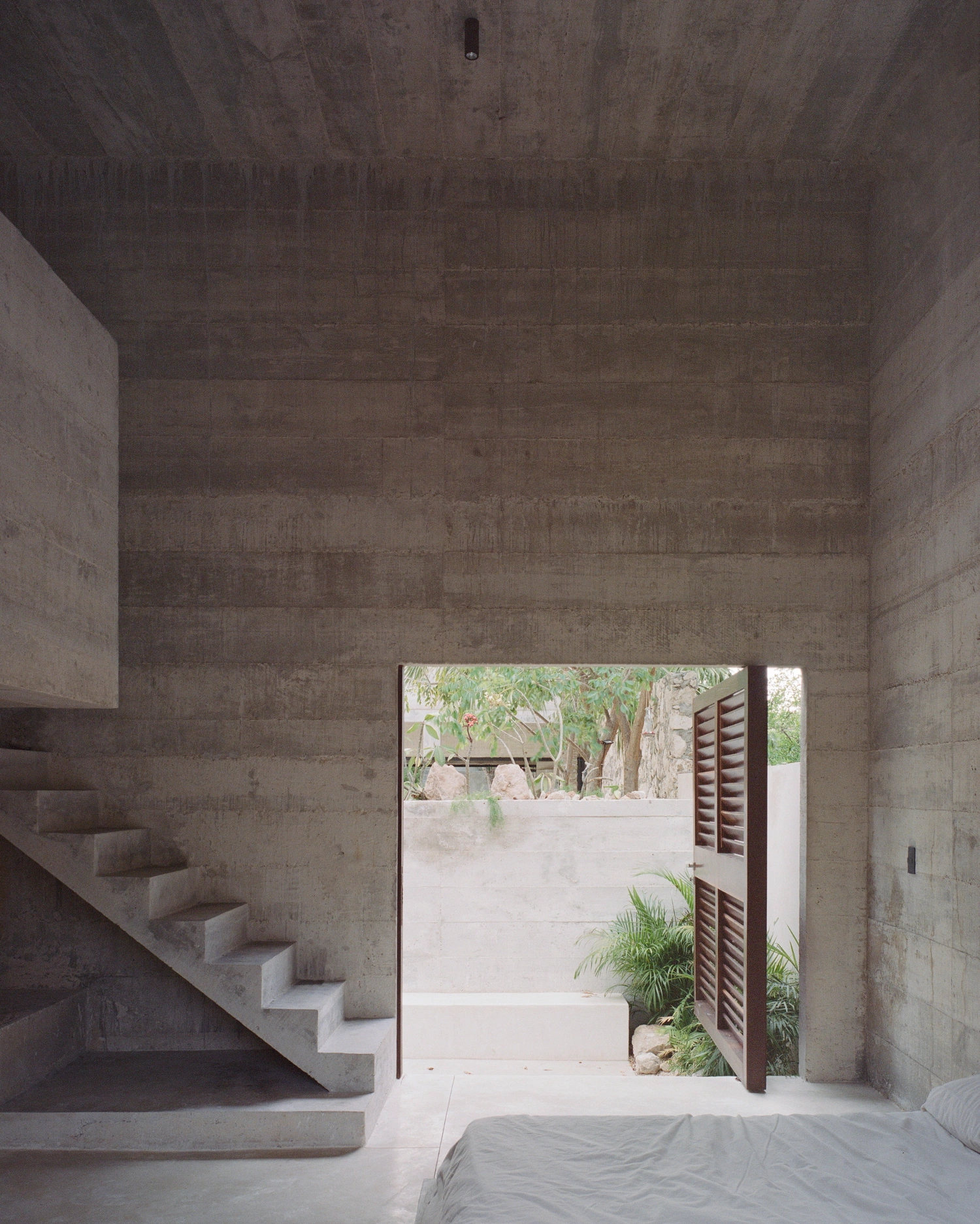 House in Los Cocos by Ludwig Godefroy Architecture — A concrete bedroom interior frames the courtyard beyond through an open timber-shuttered door.