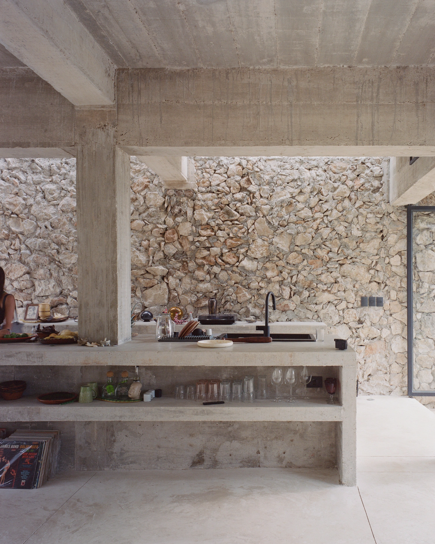 House in Los Cocos by Ludwig Godefroy Architecture — The kitchen workspace frames a dramatic stone-clad wall through the open concrete pavilion's edge.