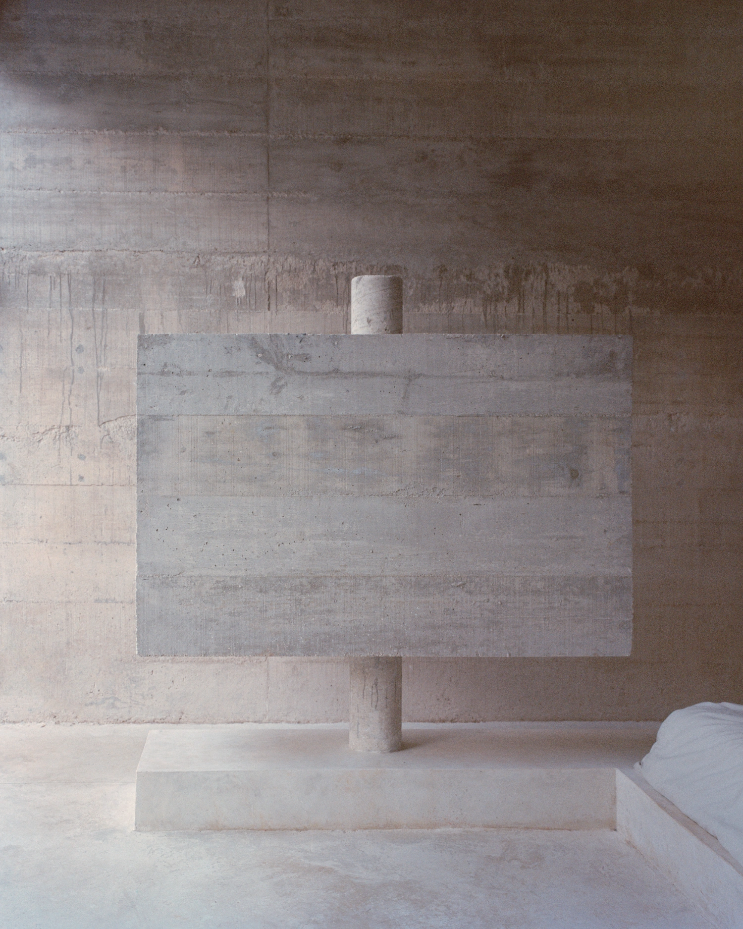 House in Los Cocos by Ludwig Godefroy Architecture — A concrete structural post rises through a rectangular stone platform, supporting a horizontal board-formed concrete panel.