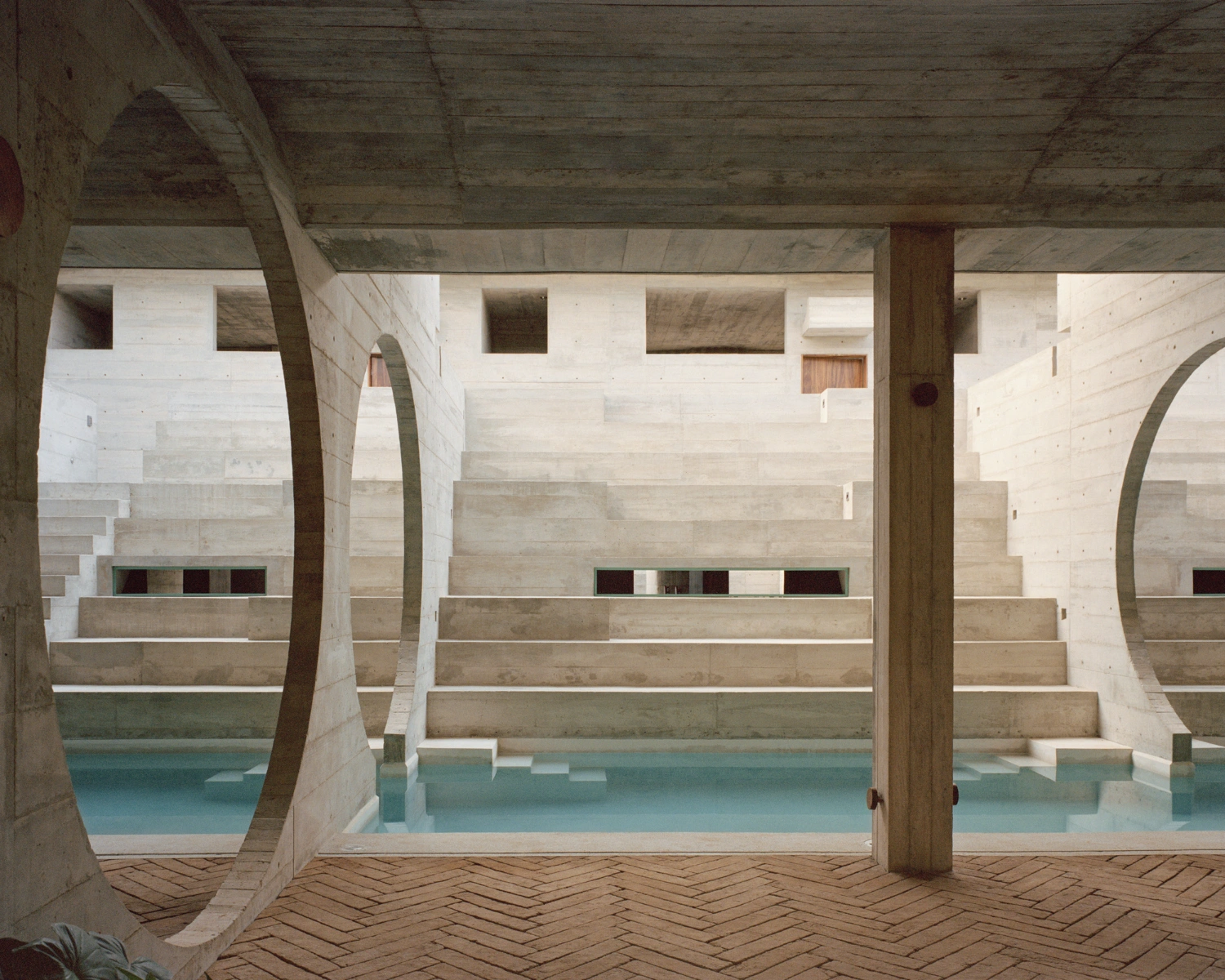 House TO by Ludwig Godefroy Architecture — A columned interior passage frames the turquoise water feature beyond, its stepped edges descending into the courtyard pool.