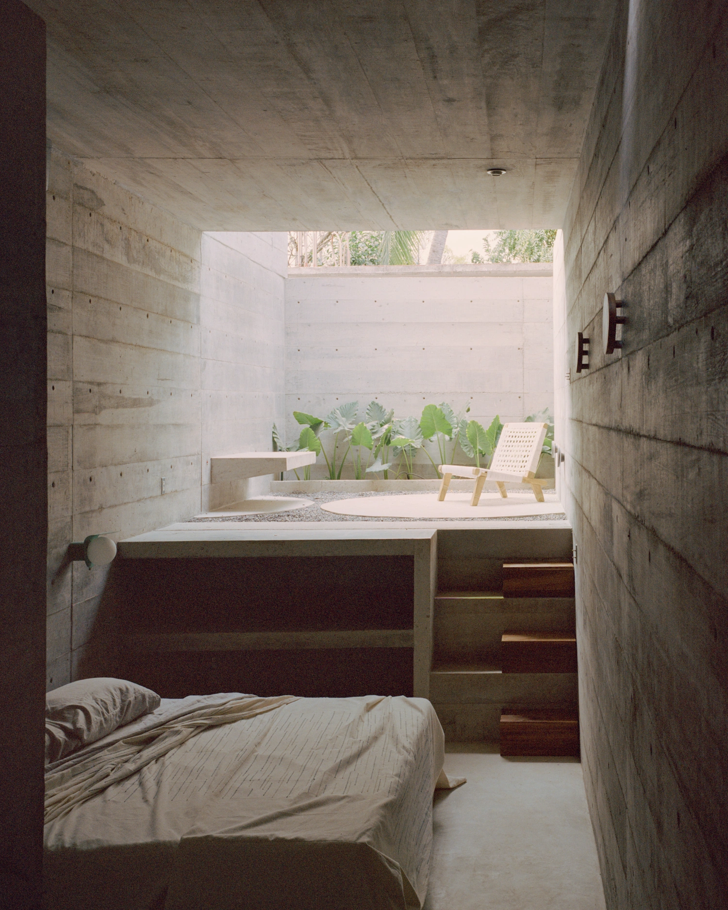 House TO by Ludwig Godefroy Architecture — A narrow bedroom chamber carved from board-formed concrete, anchored by a low platform bed with timber headboard and storage steps.