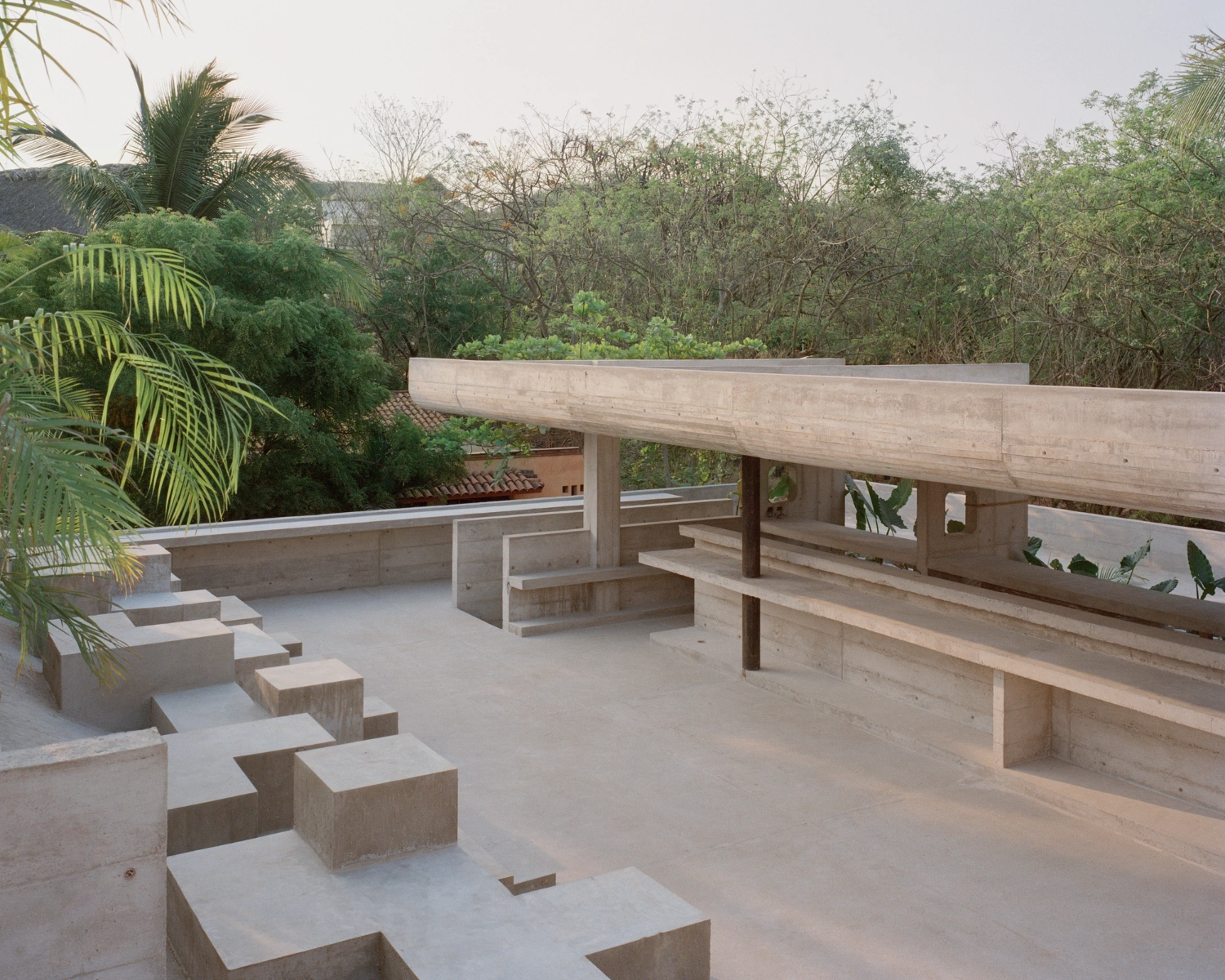 House TO by Ludwig Godefroy Architecture — A rooftop terrace rendered in board-formed concrete features stepped seating elements and horizontal bench surfaces overlooking dense tropical vegetation.