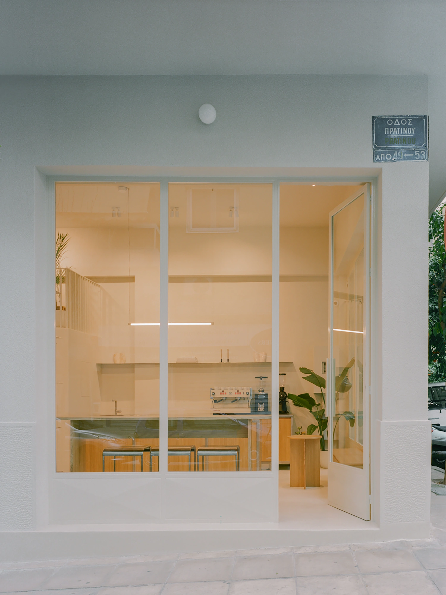 Moden Café by Georgios Apostolopoulos — Moden Café presents itself through a floor-to-ceiling glazed storefront with white aluminum framing, the glass planes interrupted by horizontal white metal...