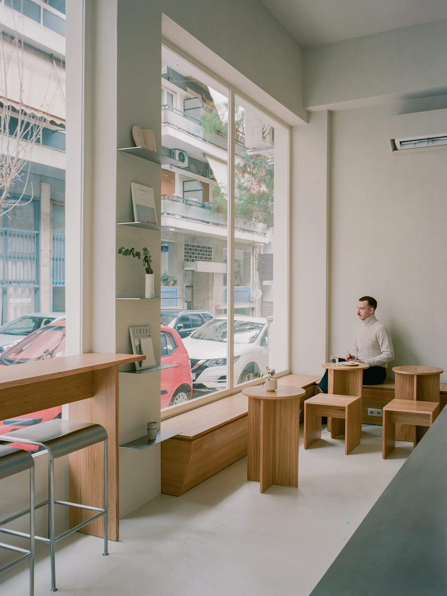 Moden Café by Georgios Apostolopoulos — Moden Café's ground-floor interior frames a street-level vantage into urban Athens through floor-to-ceiling glazing with white aluminum mullions.