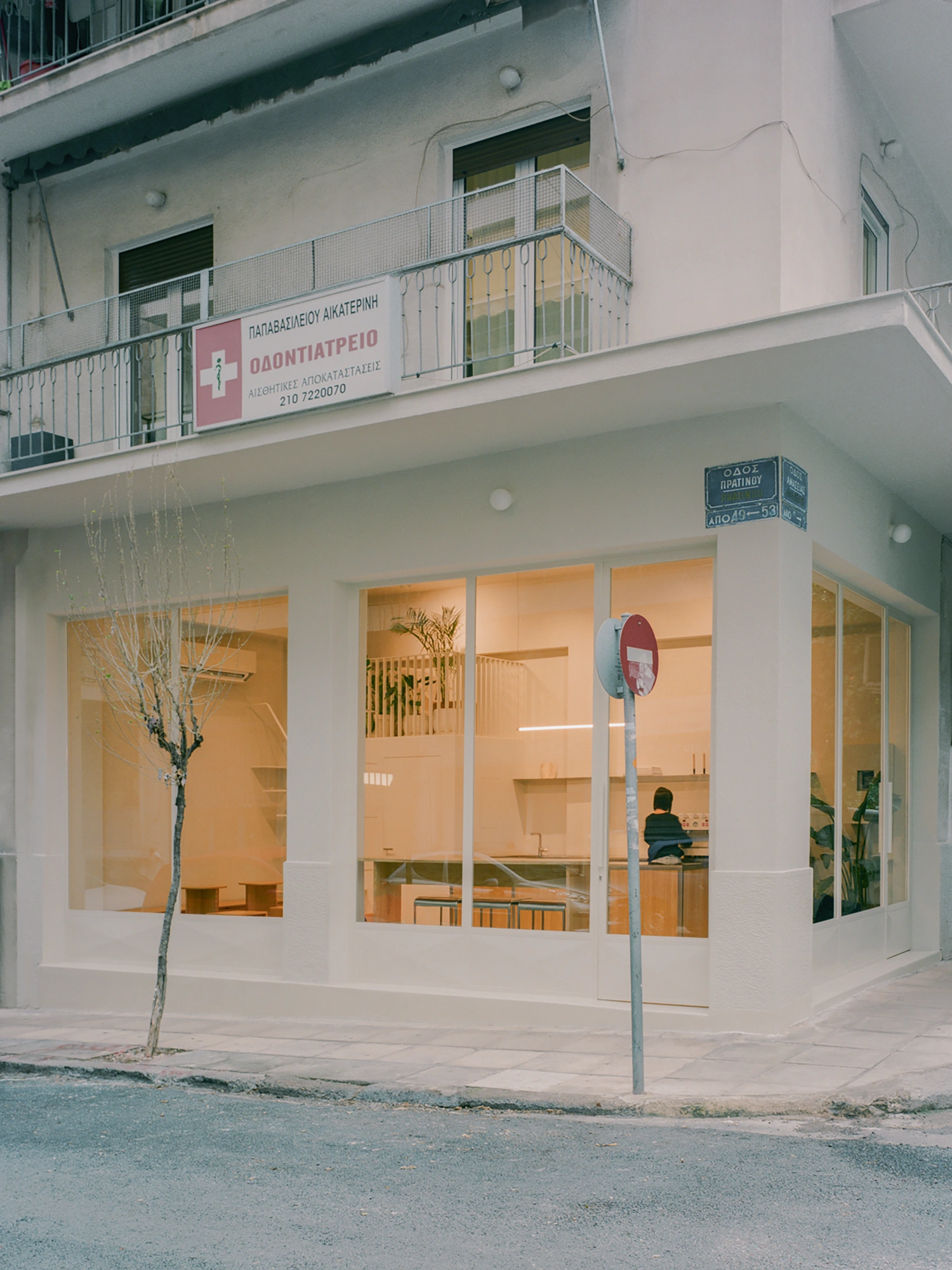 Moden Café by Georgios Apostolopoulos — The ground-floor storefront of Moden Café presents a transparent street interface: floor-to-ceiling glazing with white aluminum framing exposes the...