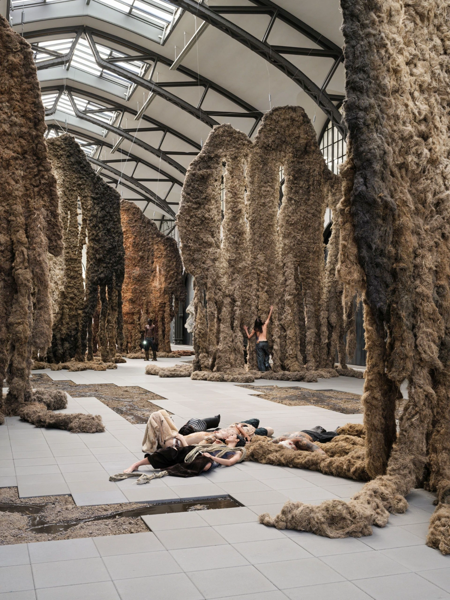 'embrace' by Klára Hosnedlová at Hamburger Bahnhof — A figure reclines on the polished concrete floor within Hosnedlová's columnar forest of woven flax tapestries.