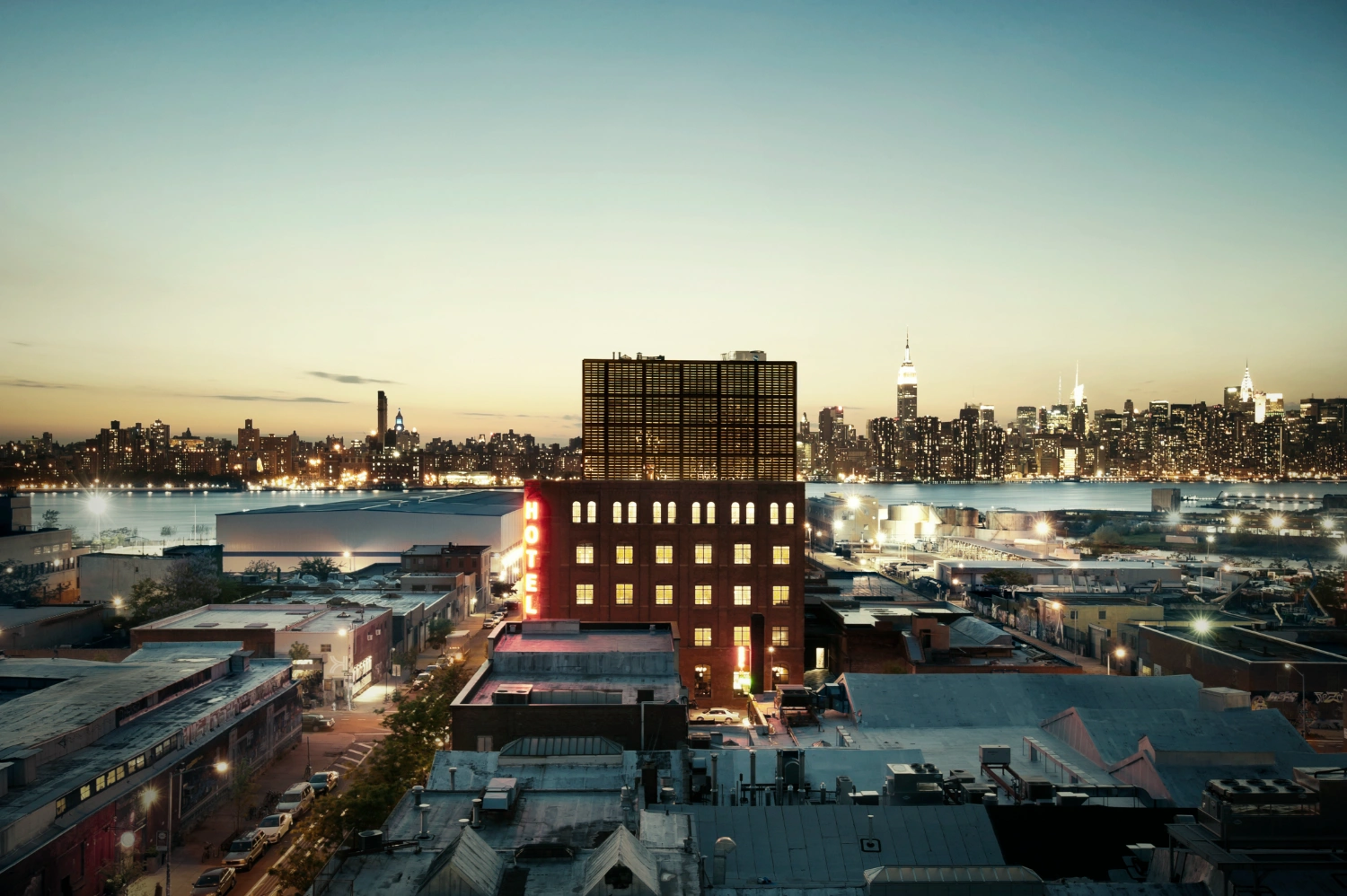 Wythe Hotel New York by Morris Adjmi Architects — Dusk photograph capturing the Wythe Hotel's red-brick facade and glass-steel penthouse against the Manhattan skyline.