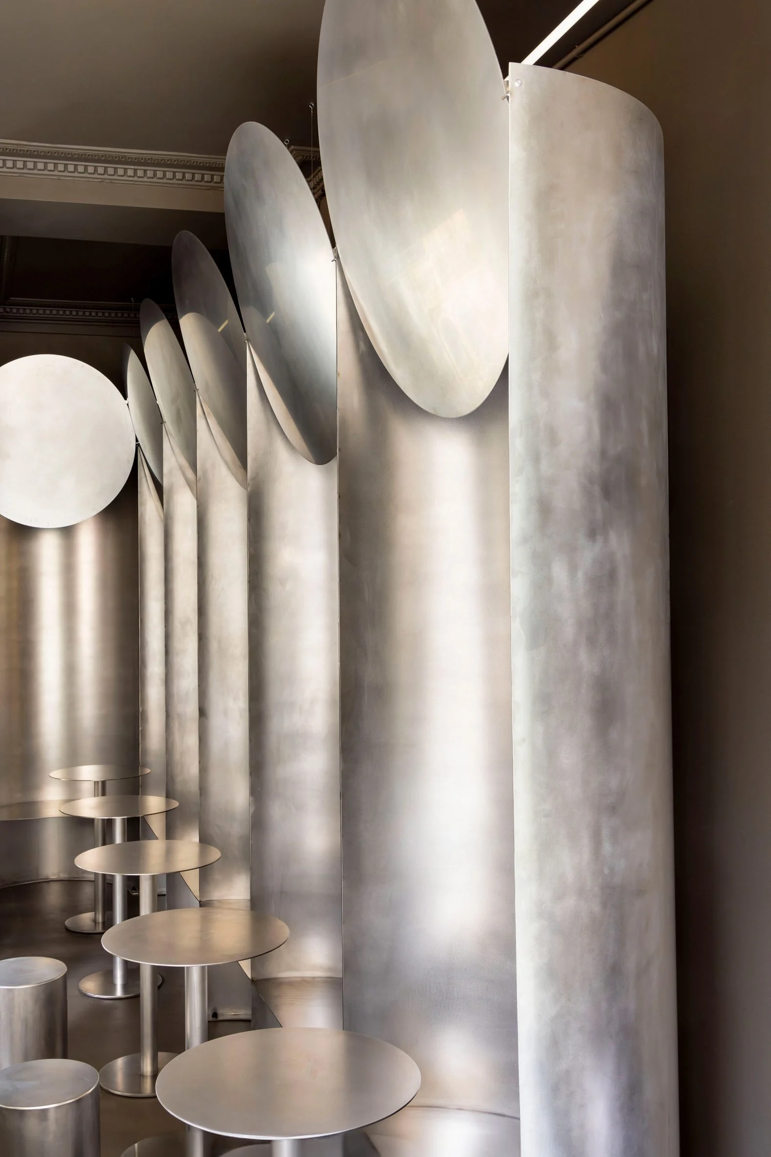 Simple Coffee by Mur Mur Architects — A row of brushed stainless steel cylindrical seating elements of graduated heights occupies the foreground, their flat circular tops catching reflected light.