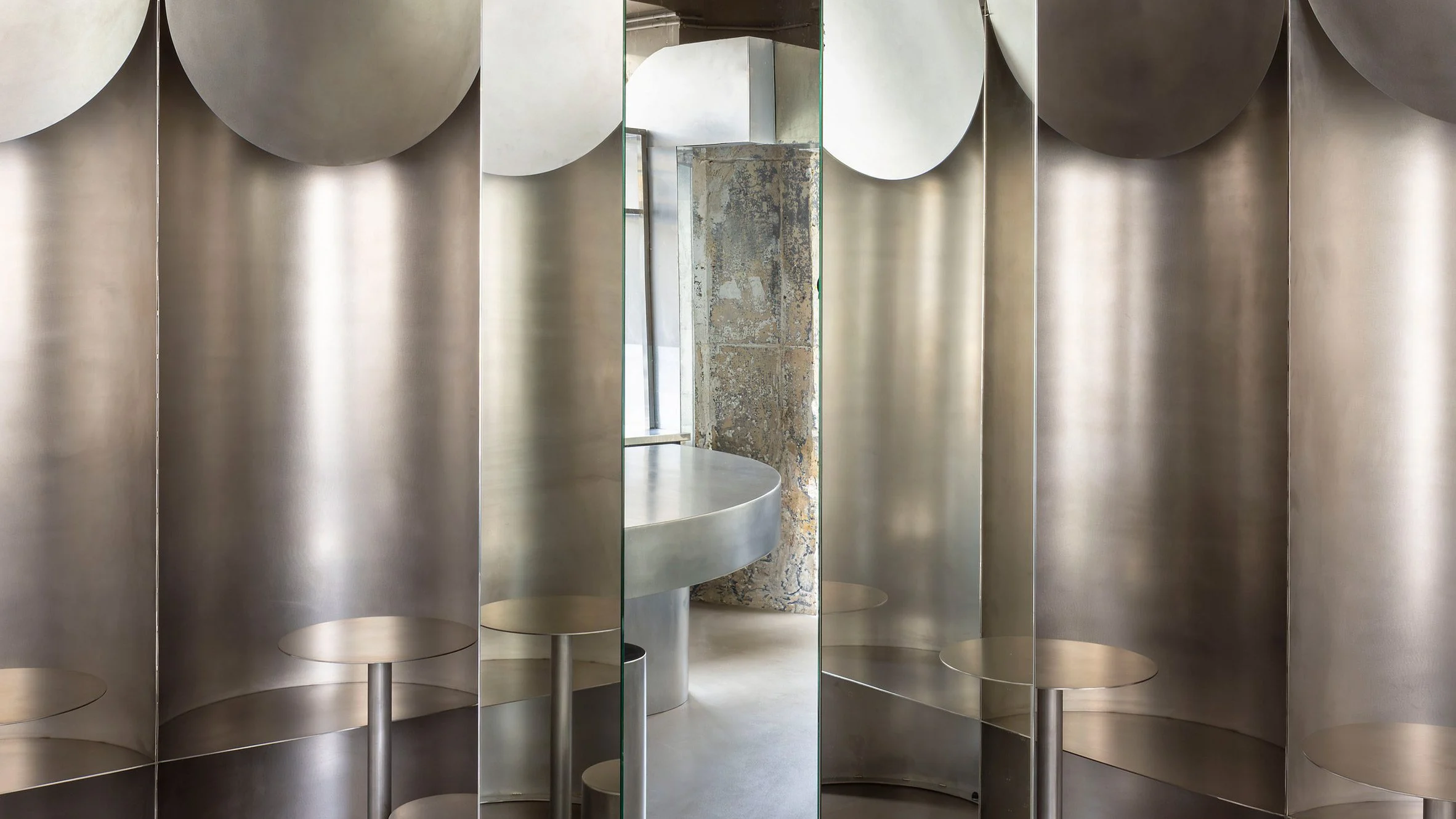 Simple Coffee by Mur Mur Architects — A single-room café interior defined by brushed stainless steel curved walls and cylindrical seating elements arranged in descending height sequence.