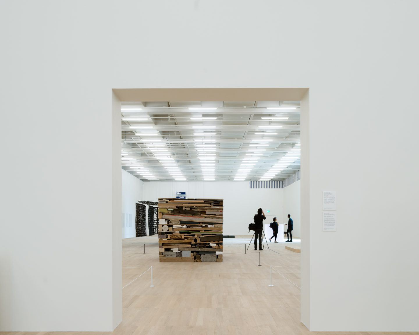 The Tate Modern by Herzog & de Meuron — A recessed gallery entrance frames a timber sculptural installation composed of stacked wooden planks in varying lengths and finishes, positioned centrally...