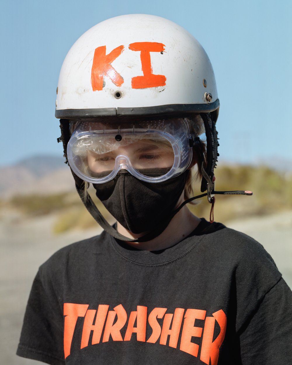 A Poor Sort of Memory by Tracy L Chandler — A child stands centered in frame wearing protective gear—white helmet marked with orange 'KI' lettering, clear safety goggles, and black face mask—against a...