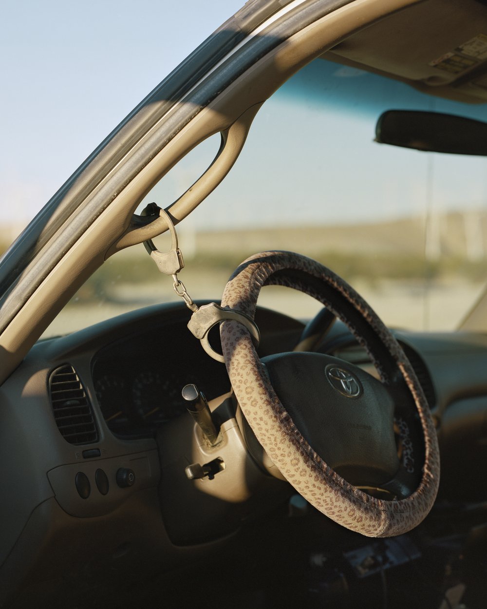 A Poor Sort of Memory by Tracy L Chandler — A Toyota steering wheel wrapped in textured fabric sits abandoned in the cab of a vehicle parked in the California Desert.