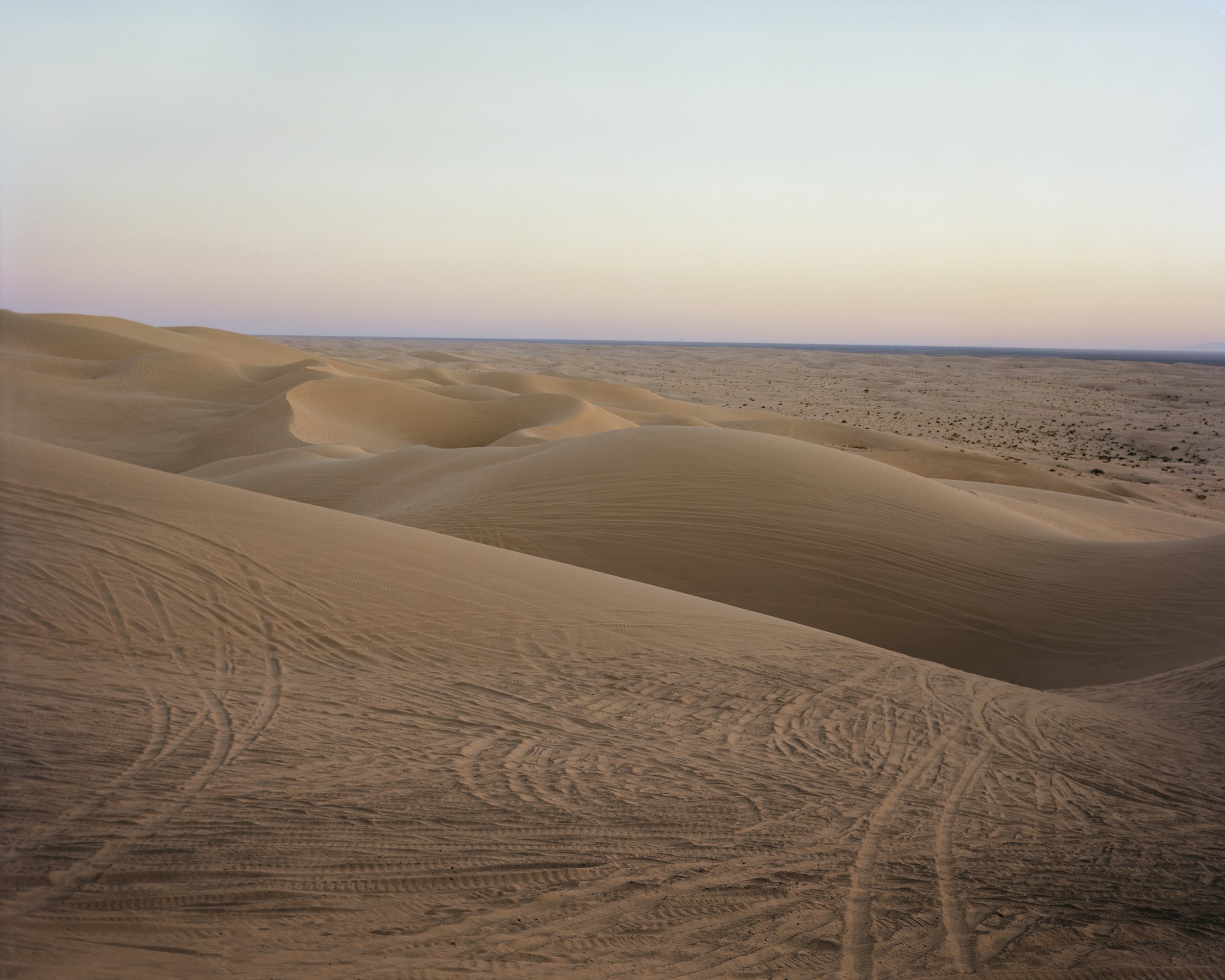 A Poor Sort of Memory by Tracy L Chandler — Wind-carved sand dunes dominate the foreground, their surfaces etched with linear ripple patterns from vehicle tracks that scar the pale tan surface.