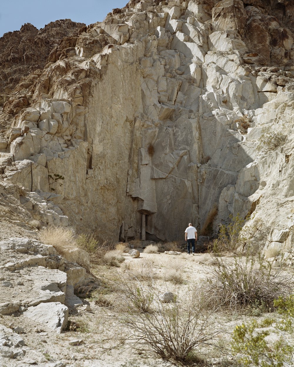 A Poor Sort of Memory by Tracy L Chandler — A solitary figure in white stands before a massive carved stone face embedded in a desert hillside, dwarfed by the monumental scale of the rock formation.