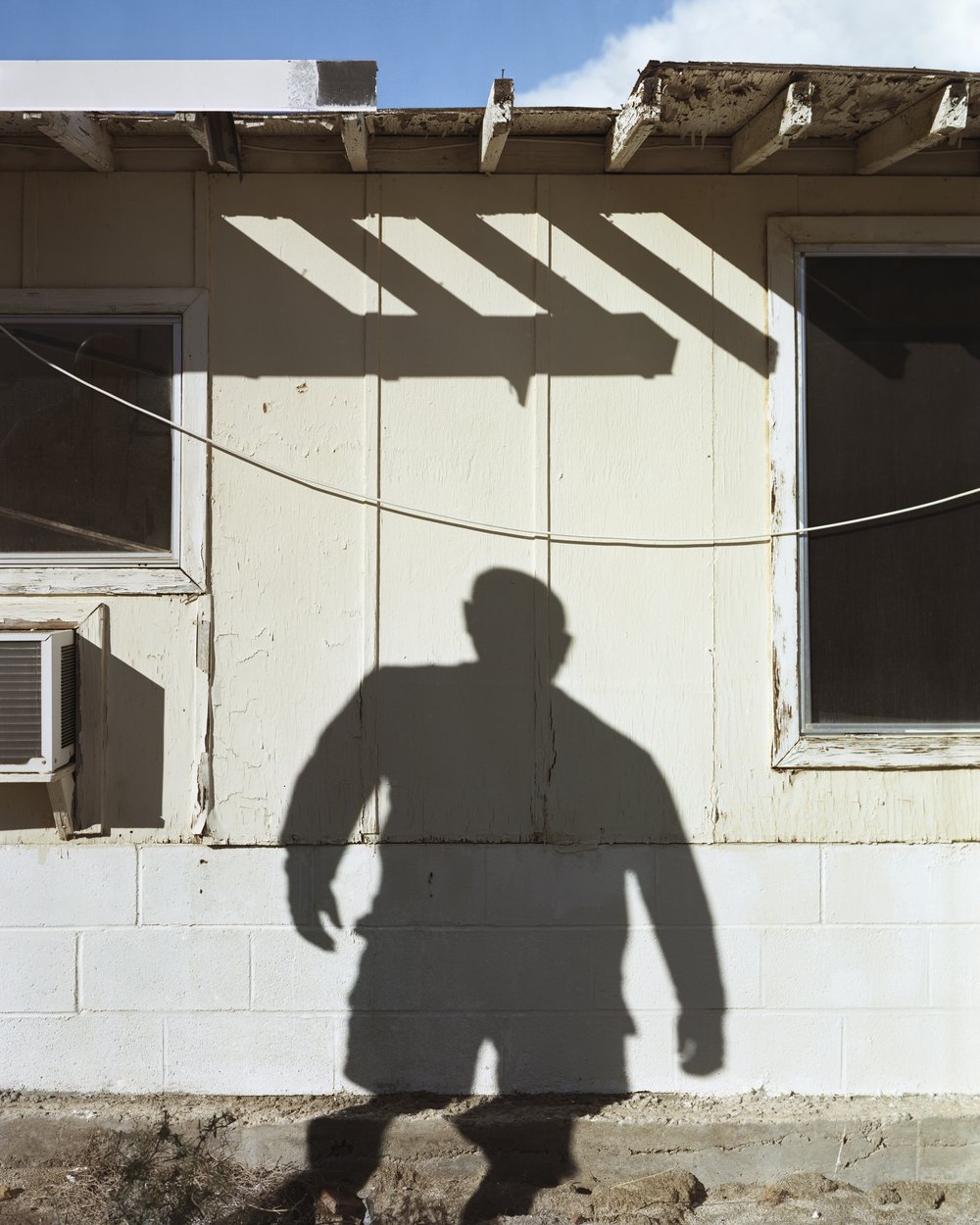 A Poor Sort of Memory by Tracy L Chandler — A figure casts a sharp silhouette against a weathered white cinder-block wall, their shadow bisected by diagonal bands of sunlight filtering through a...