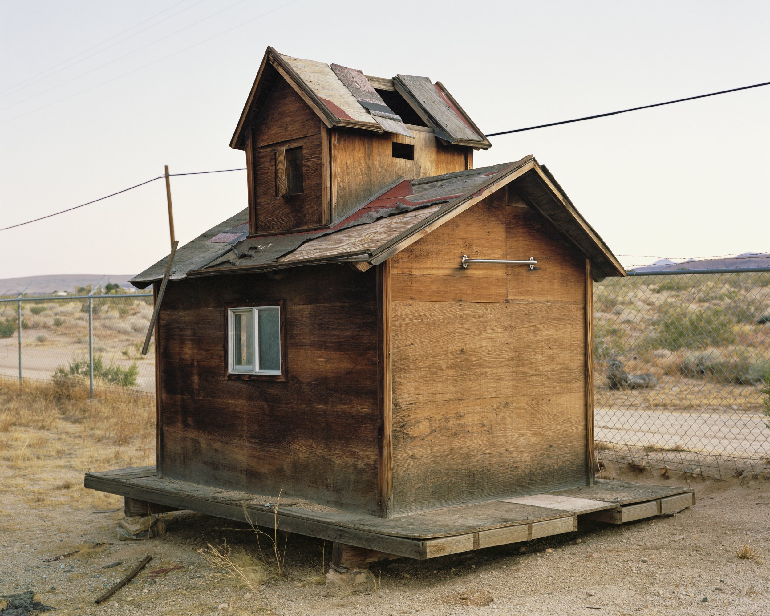 A Poor Sort of Memory by Tracy L Chandler — A timber-frame structure with pitched roof and central tower element sits elevated on a wooden platform in the California desert.