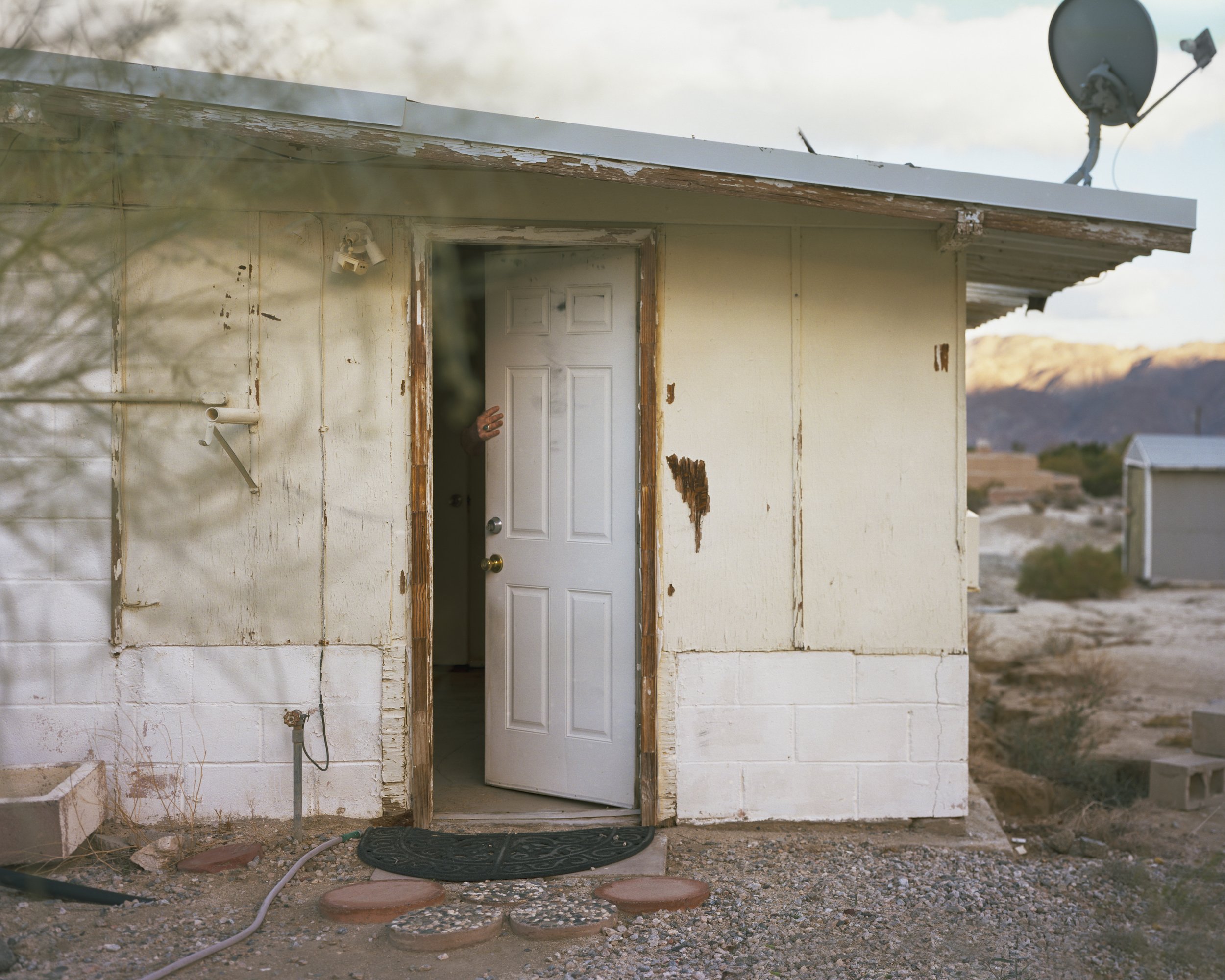 A Poor Sort of Memory by Tracy L Chandler — A deteriorating single-wide trailer with white painted wood siding and exposed timber framing around the entrance door sits isolated in the California desert.