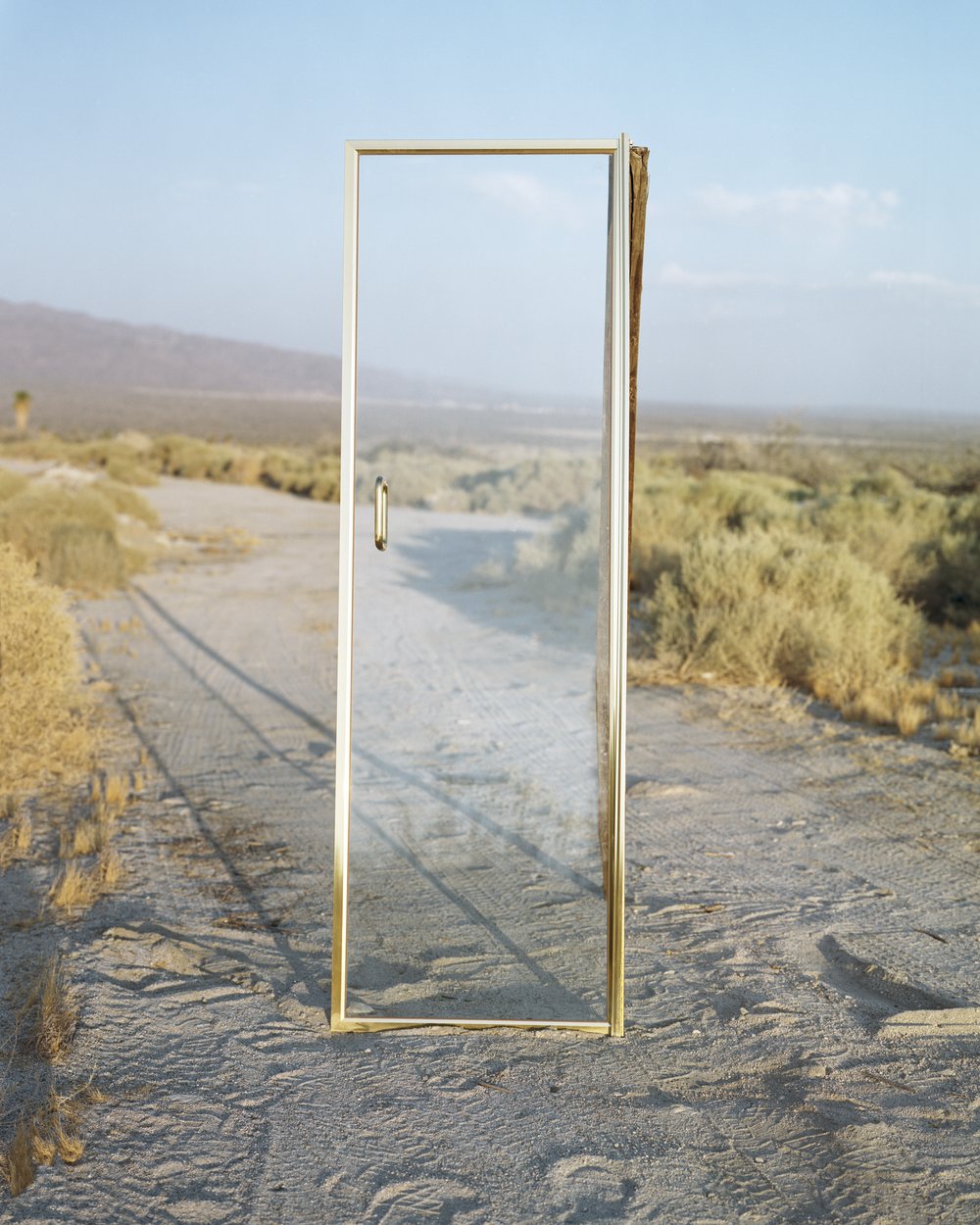 A Poor Sort of Memory by Tracy L Chandler — A weathered brass-framed glass door stands isolated on packed desert floor, its transparent panes reflecting the arid landscape beyond.