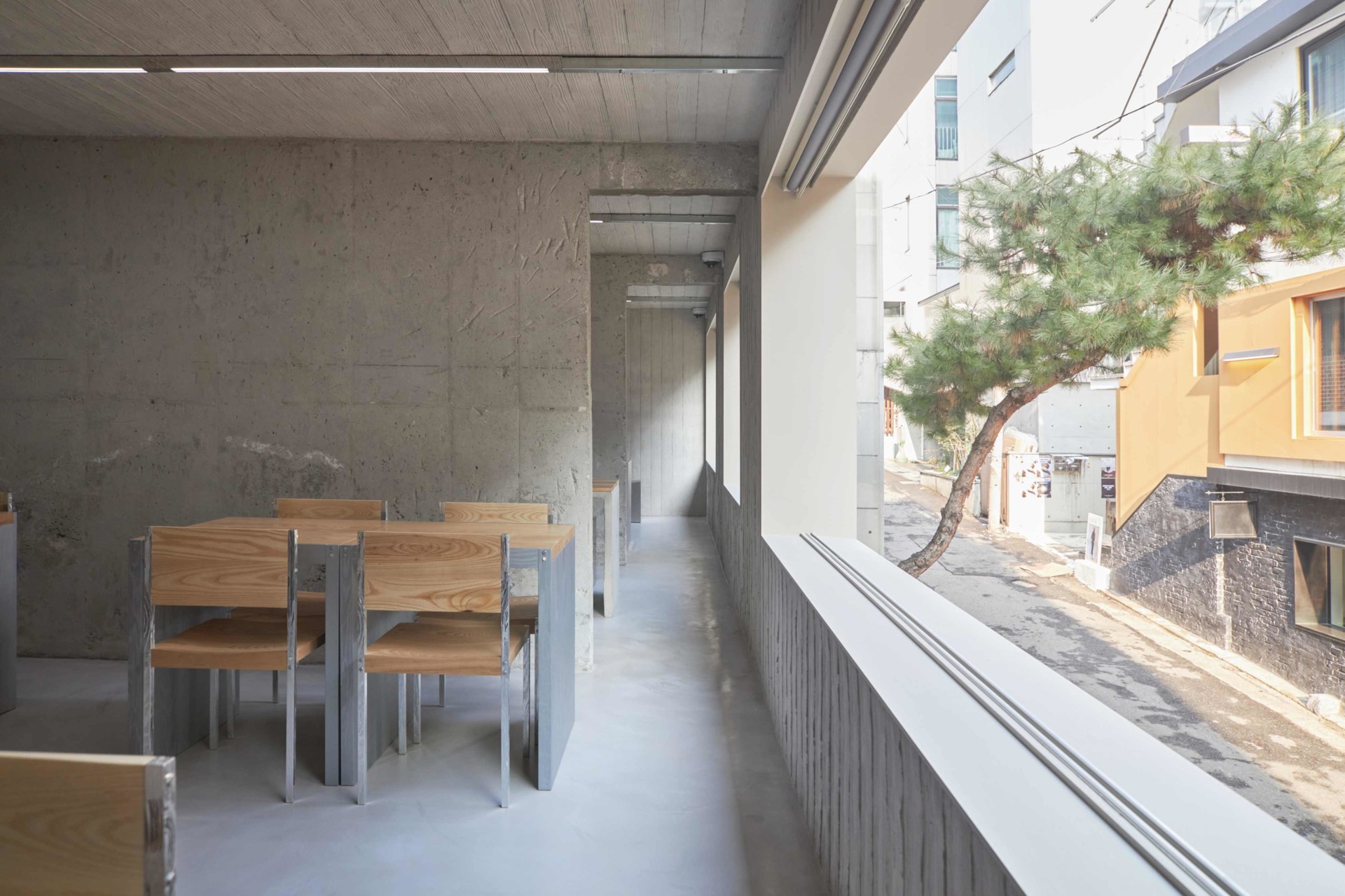 HAHOUSE Cafe by Niceworkshop — A narrow corridor unfolds with a dining table and chairs positioned along the left wall, their timber seats and stainless steel frames anchoring the passage.
