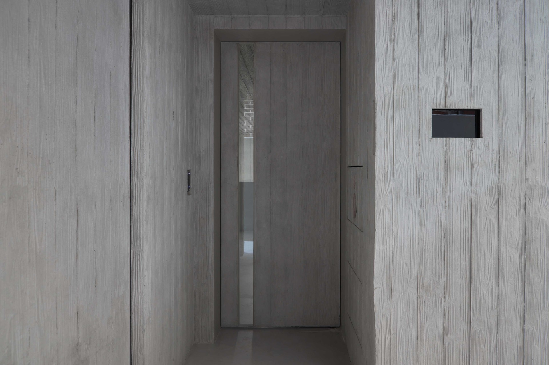 HAHOUSE Cafe by Niceworkshop — A narrow corridor entrance frames a dark timber door with a vertical glass panel, its wood grain visible through the translucent pane.