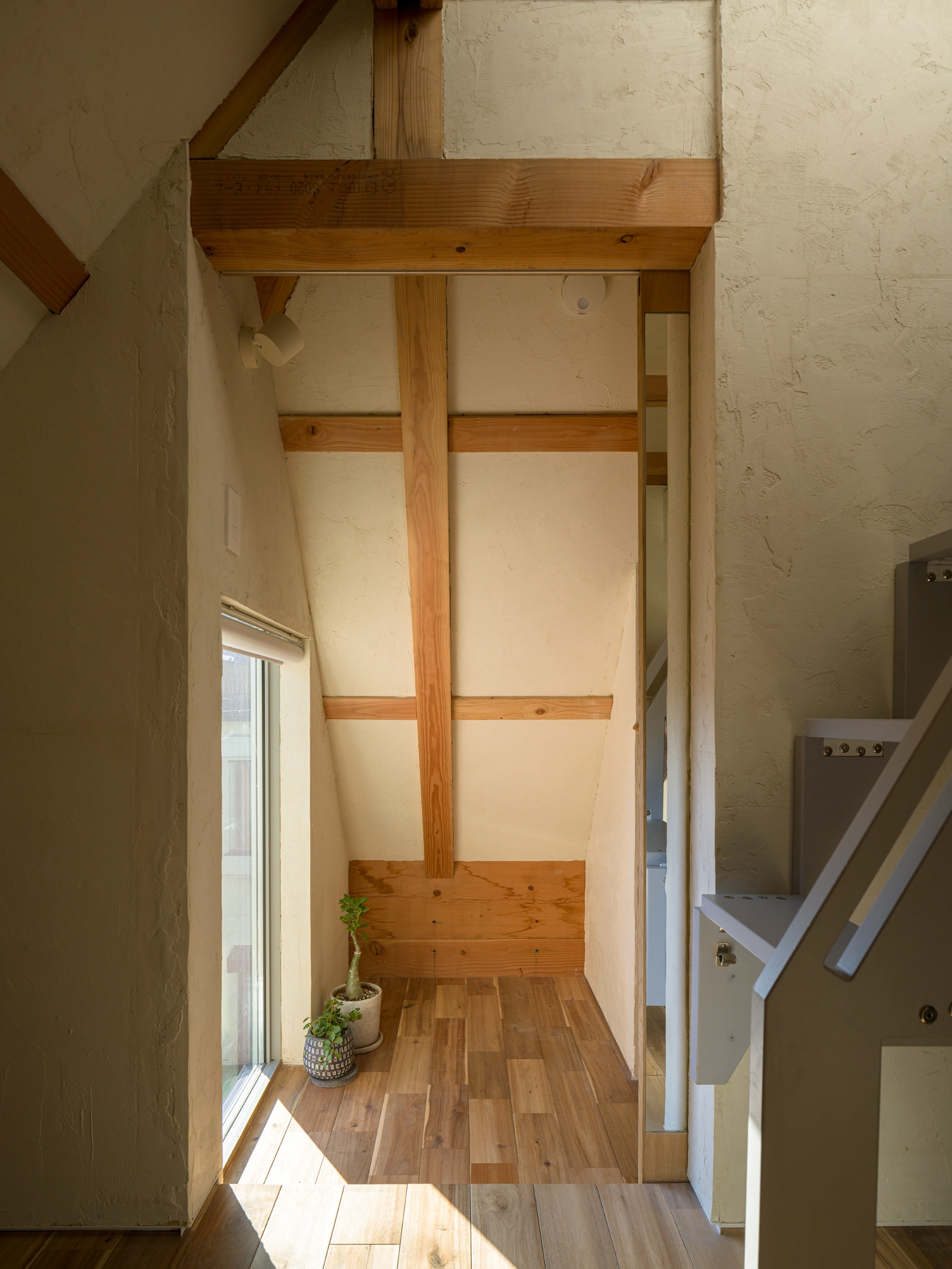Small House on a Corner Lot by KOMINORU Design — A narrow interior corridor defined by post-and-beam timber framing, where vertical cedar posts and horizontal beams segment cream-colored plaster walls into...