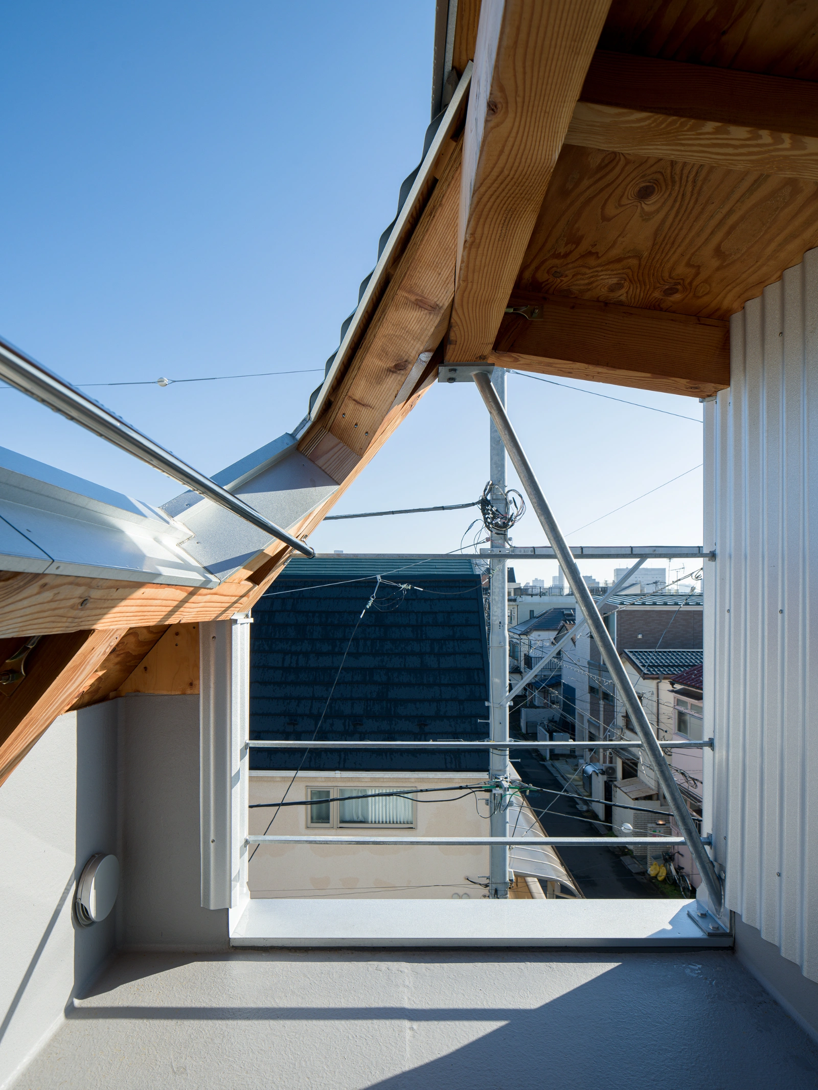 Small House on a Corner Lot by KOMINORU Design — A rooftop terrace viewed from beneath a timber-framed overhang, its angled soffit exposing raw wood grain and knots.