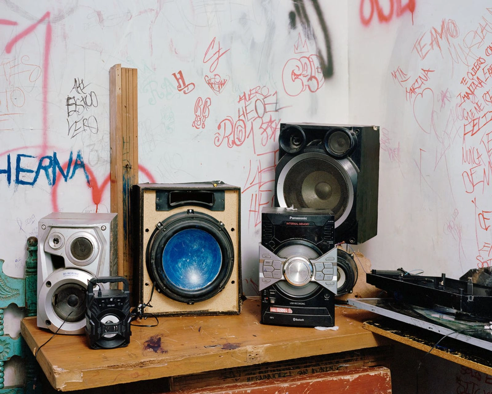Bravo by Felipe Romero Beltrán — A wooden table laden with obsolete audio equipment occupies the foreground of this composition: a cream-coloured speaker cabinet with exposed blue-painted...
