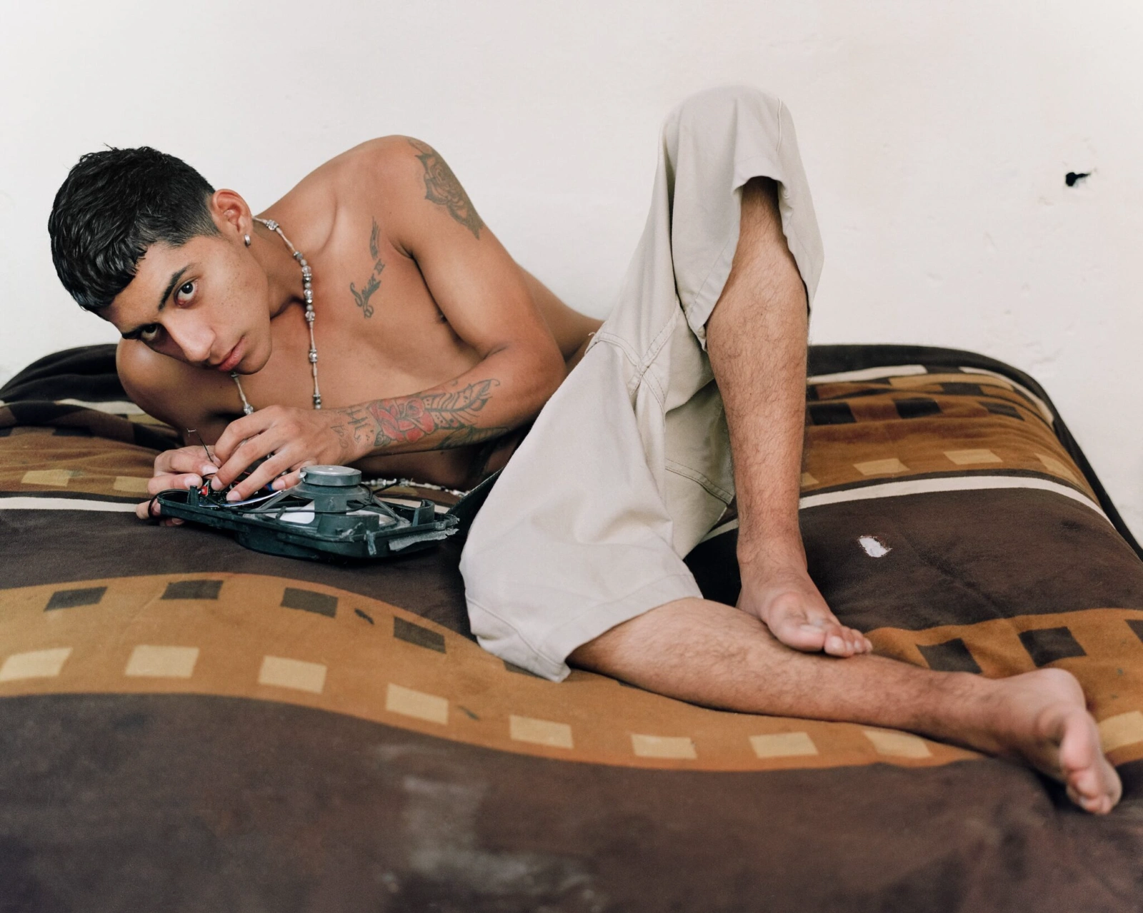 Bravo by Felipe Romero Beltrán — A shirtless figure reclines on a patterned blanket across a bed, torso angled toward the camera with a beaded chain necklace and visible tattoos on...