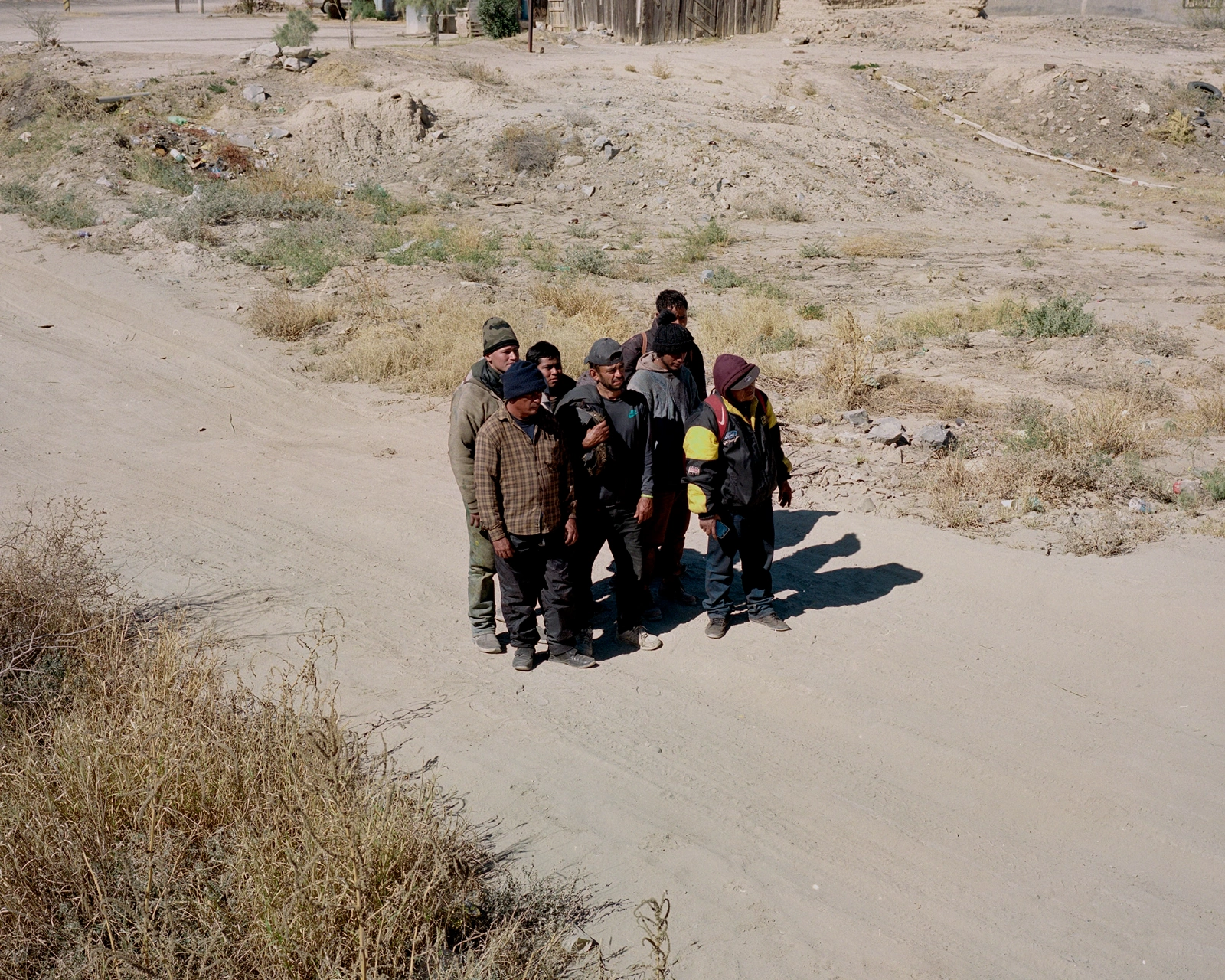 Bravo by Felipe Romero Beltrán — A group of seven figures gathered on bare earth in Rio Bravo, their bodies clustered tightly in the center of an expansive, largely vacant lot.