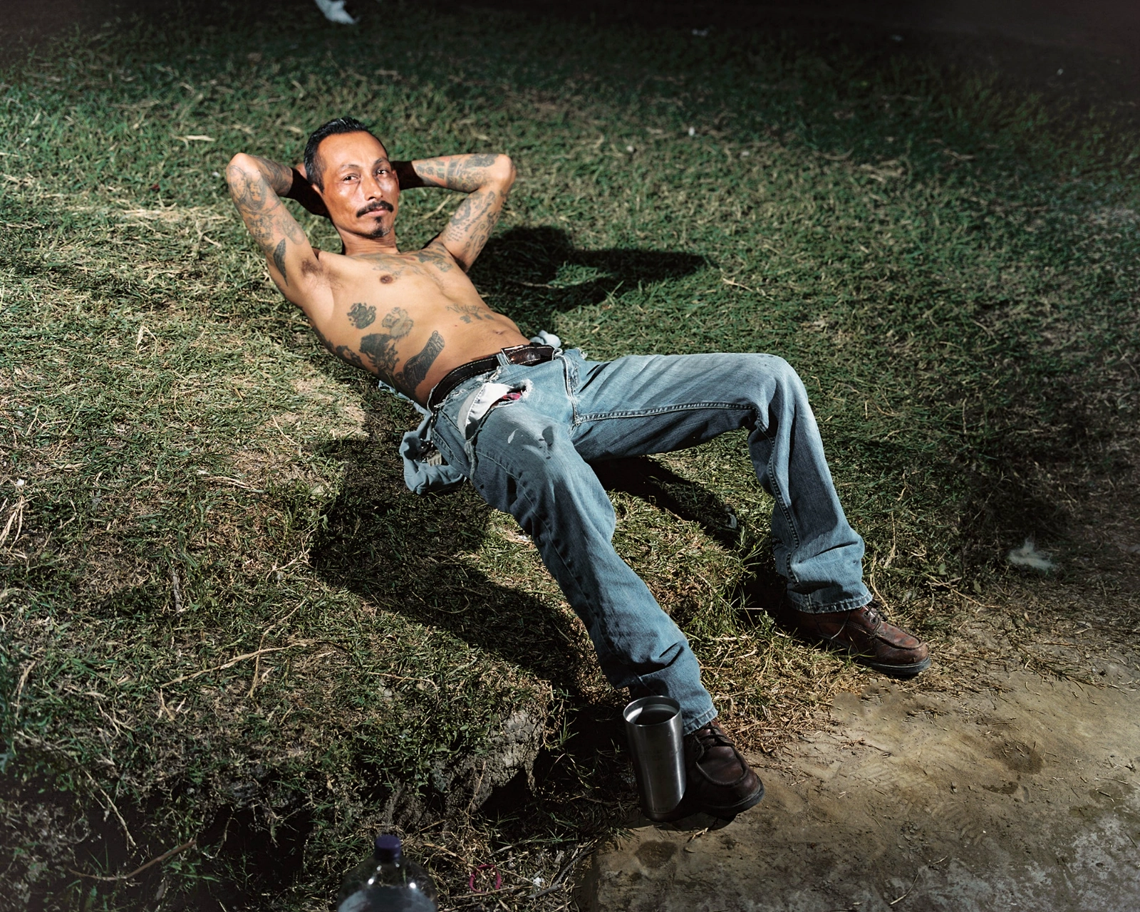 Bravo by Felipe Romero Beltrán — A shirtless man with multiple tattoos reclines on dry grass and packed earth, his body angled across the frame in a posture of repose or exhaustion.
