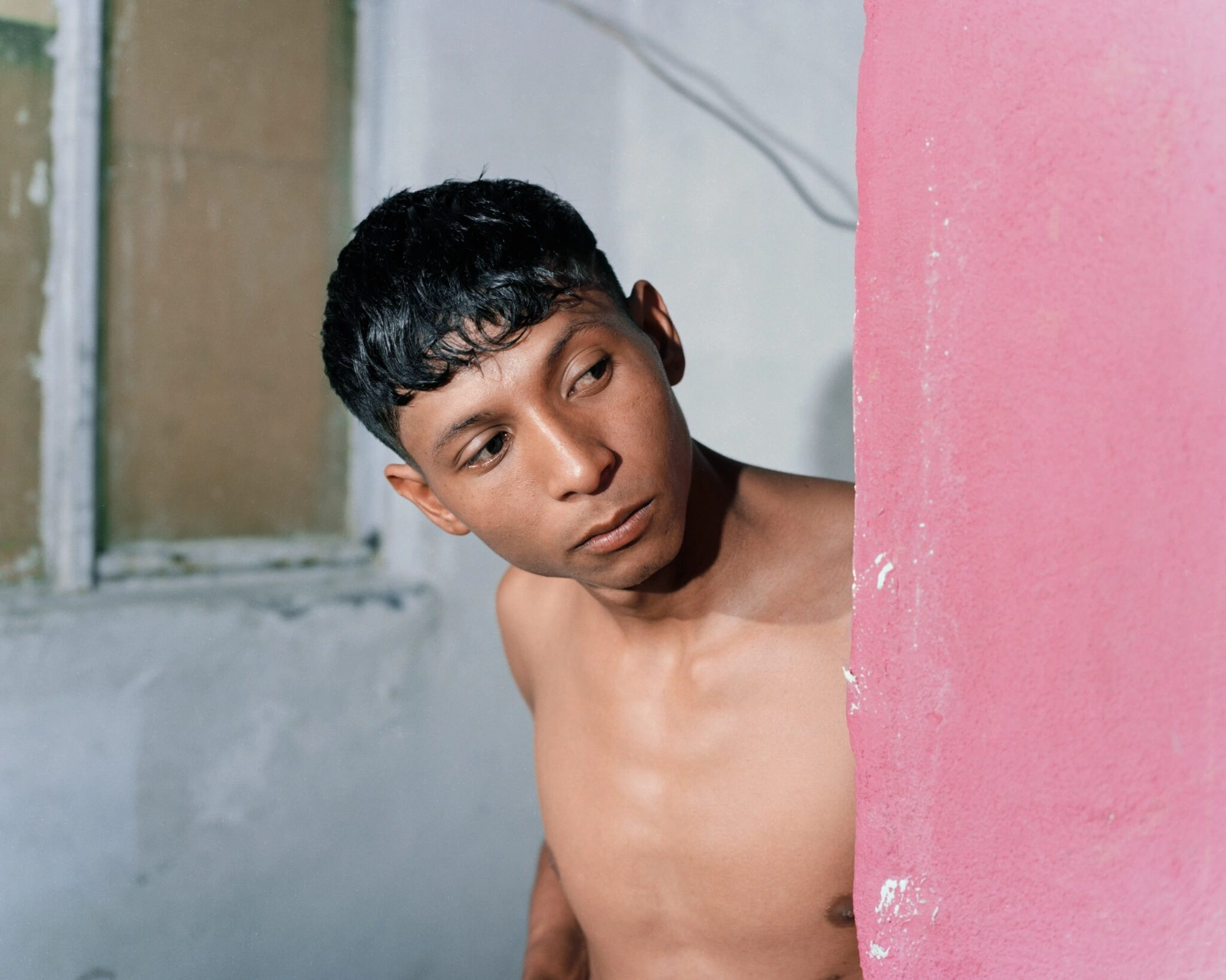 Bravo by Felipe Romero Beltrán — A shirtless subject positioned at the corner junction of a two-tone rendered wall—terracotta-red on the right, slate-grey on the left—peers toward the...