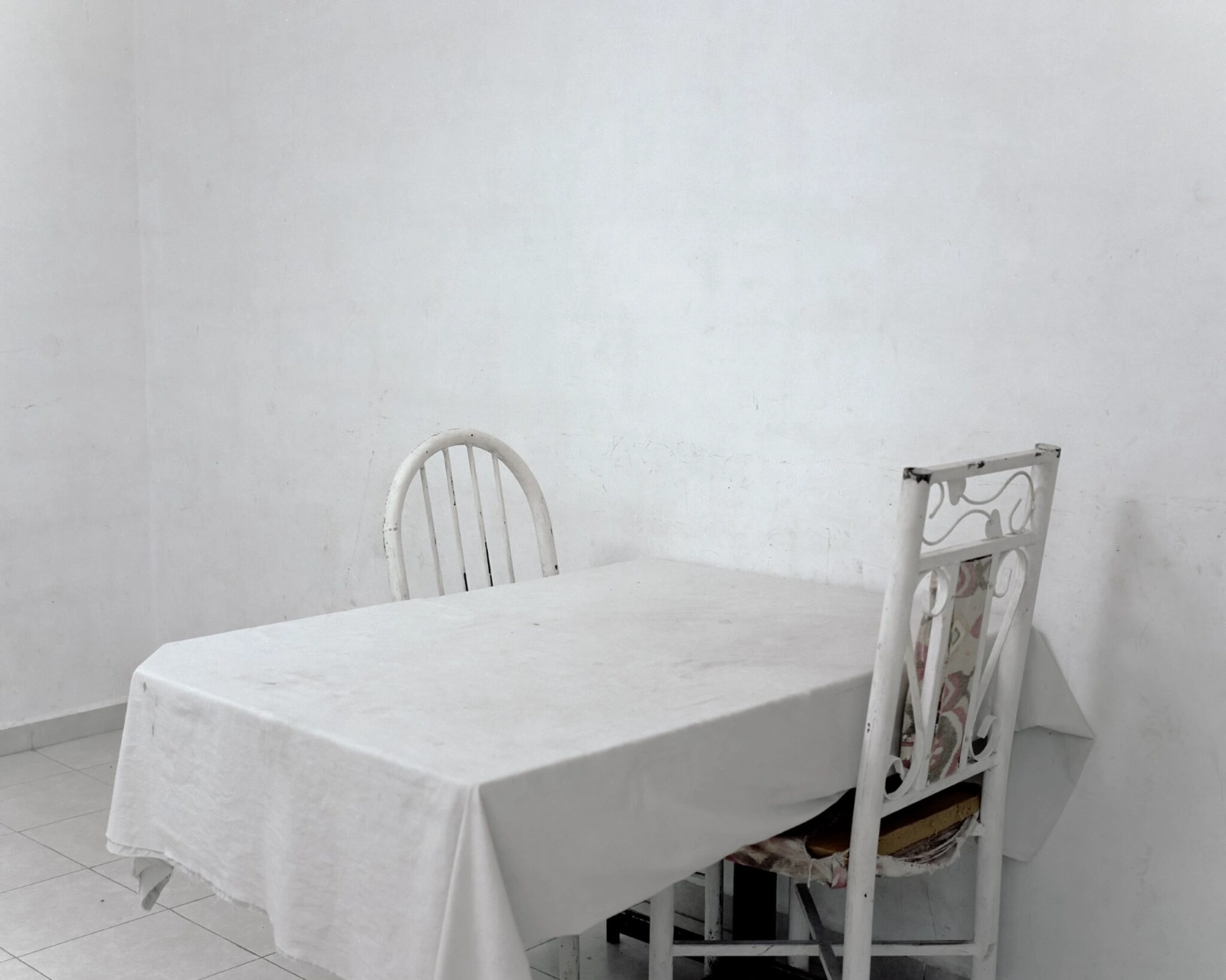 Bravo by Felipe Romero Beltrán — A sparse domestic interior in Rio Bravo presents an oval table draped in white linen and two metal chairs—one with vertical spindle back, the other ornate...