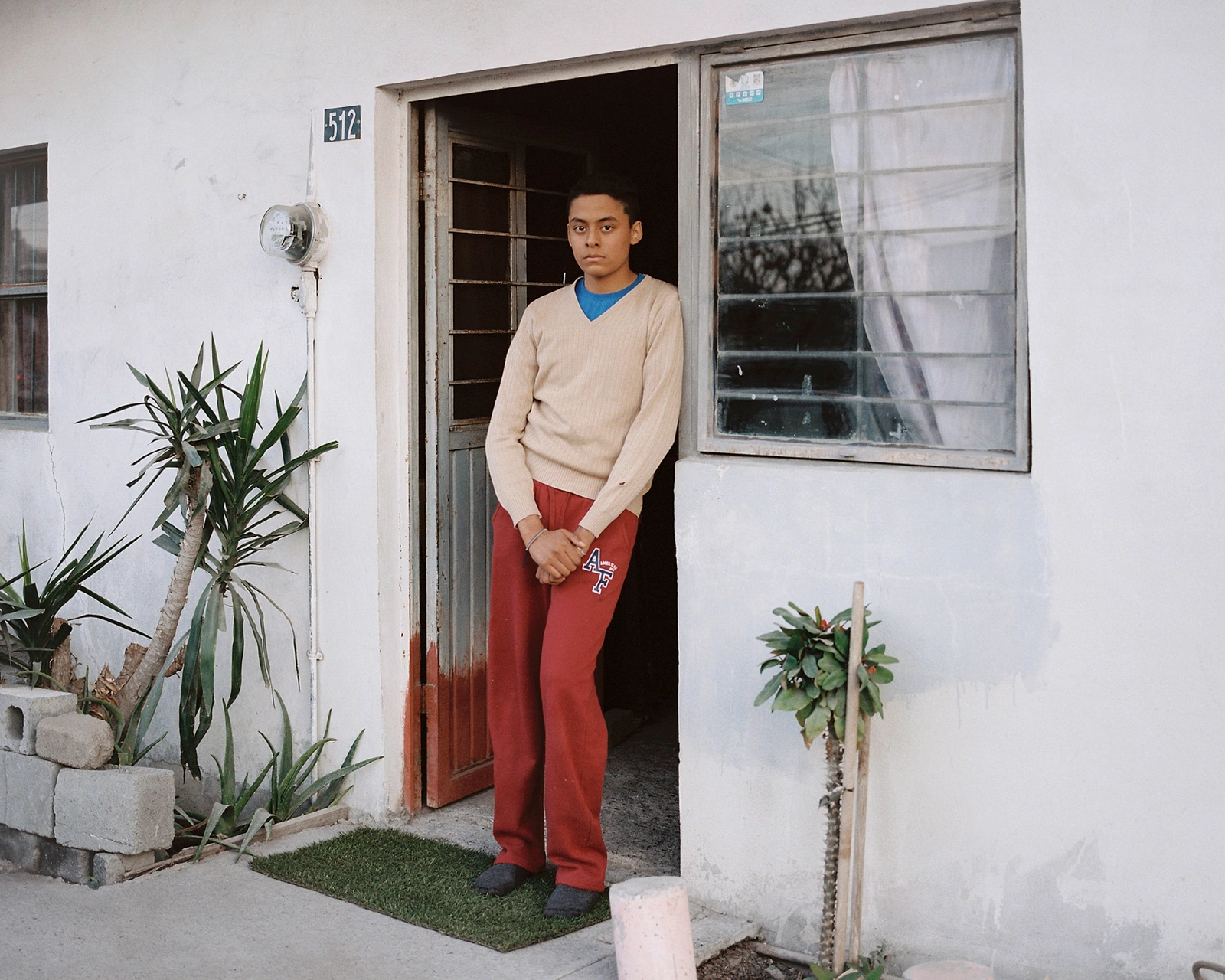 Bravo by Felipe Romero Beltrán — A portrait of a young man standing in the doorway of a modest concrete dwelling at number 512, framed by the sharp geometric division between...