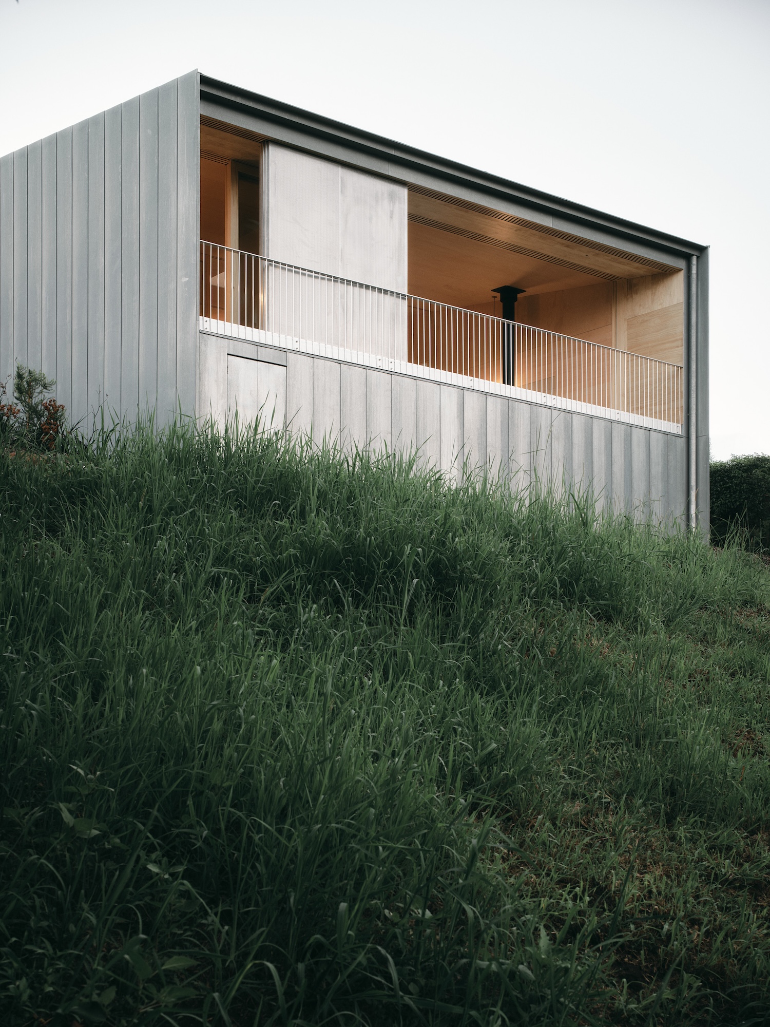 Mapleton House by Atelier Chen Hung — The exterior facade presents a monolithic rendered wall in pale grey-white, vertically ribbed with regular corrugations that catch shadow and define the...