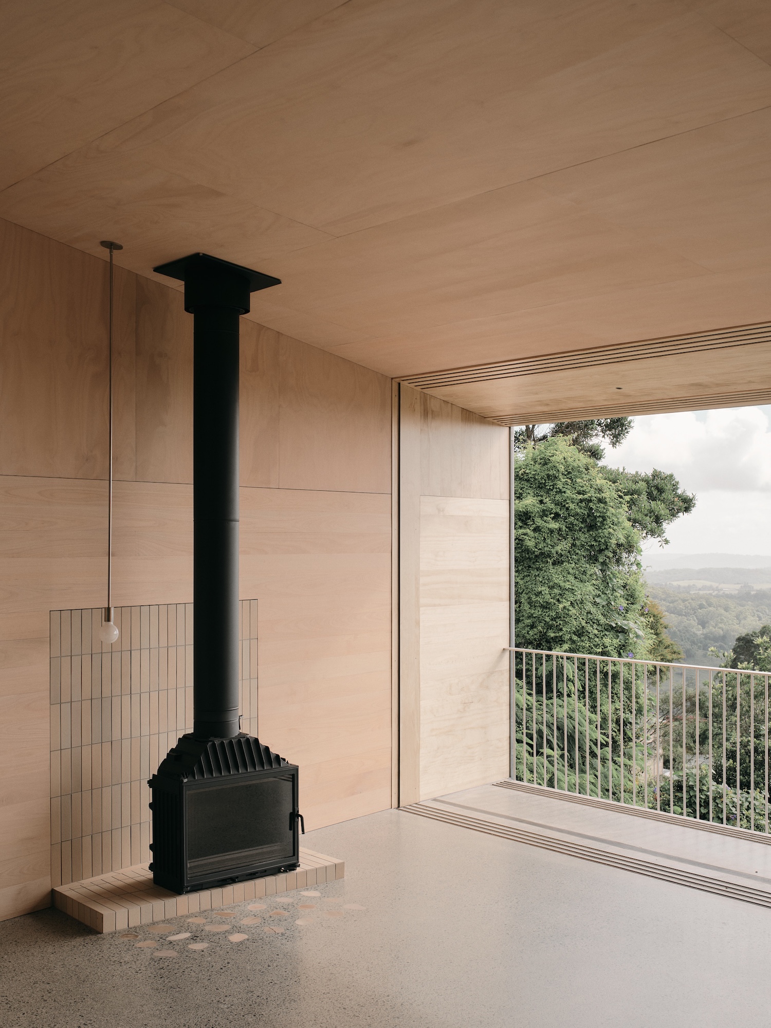 Mapleton House by Atelier Chen Hung — A monolithic black wood-burning stove anchors the open-plan interior, its tall chimney rising to meet the timber ceiling.