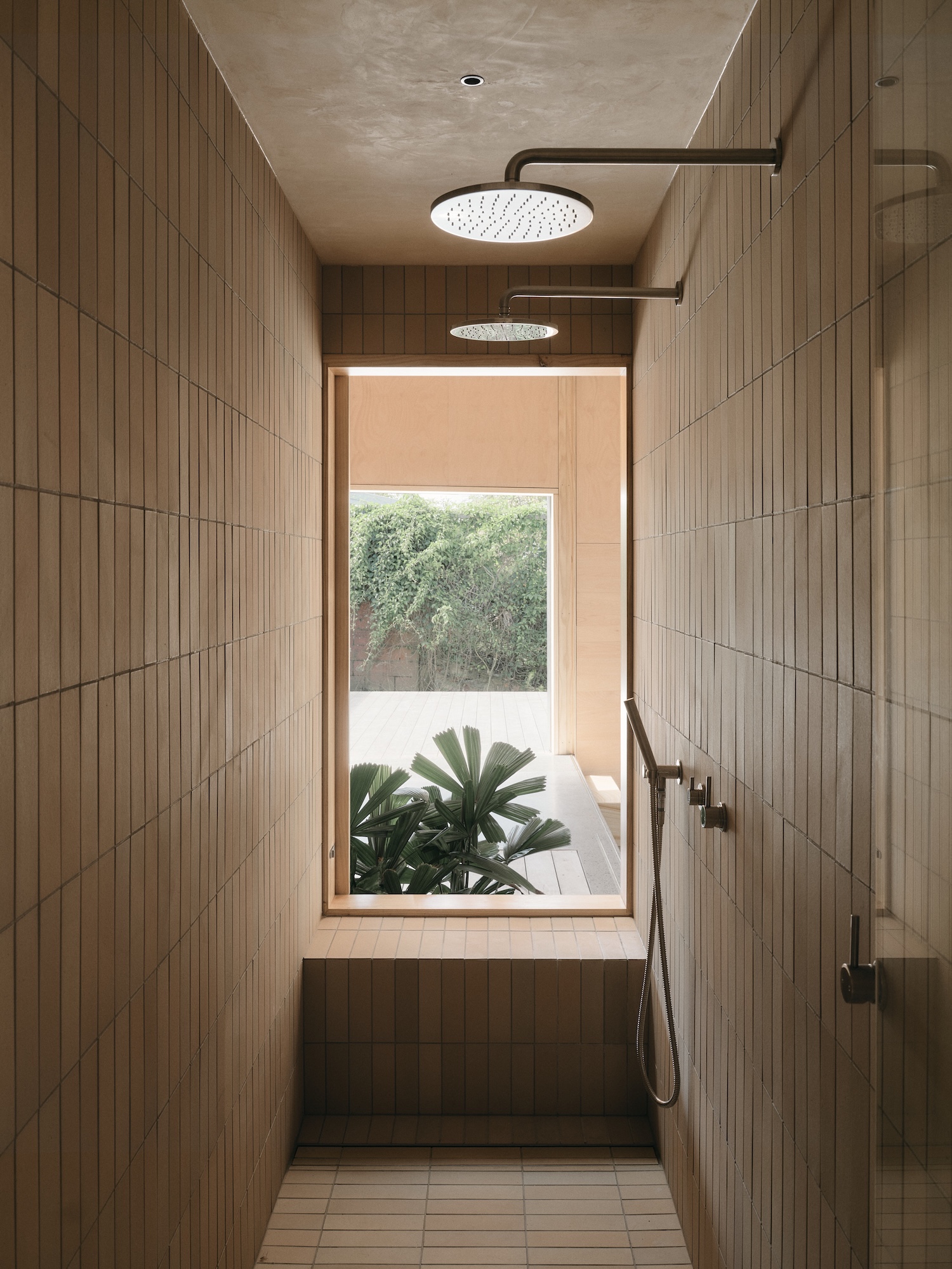 Mapleton House by Atelier Chen Hung — A corridor-like shower space lined with vertically-stacked ceramic tile in warm beige tones, terminated by a large rectangular window framing vegetation and...