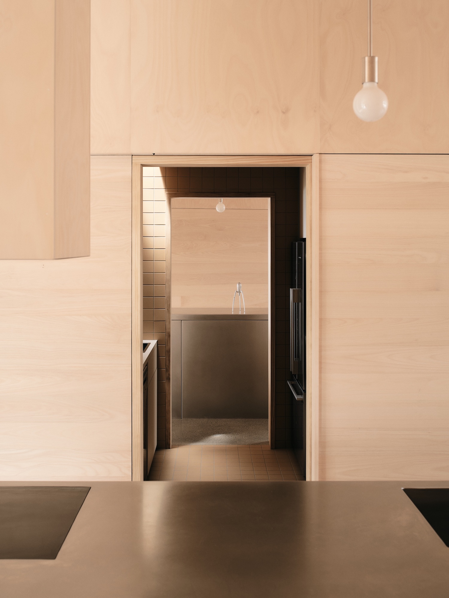 Mapleton House by Atelier Chen Hung — A view through a timber-framed aperture reveals a kitchen volume defined by monolithic rendered walls in warm ochre tones.