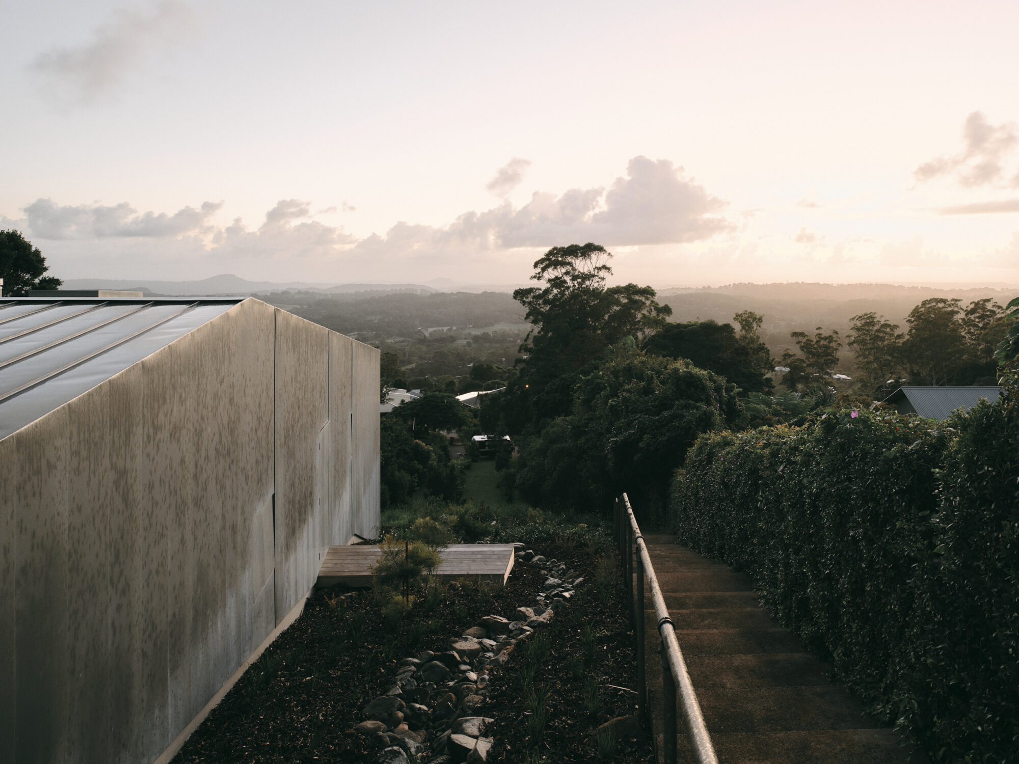 Mapleton House by Atelier Chen Hung — From the elevated vantage of the rear garden pathway, the rendered exterior wall of Mapleton House appears as a monolithic cream-colored plane, its...
